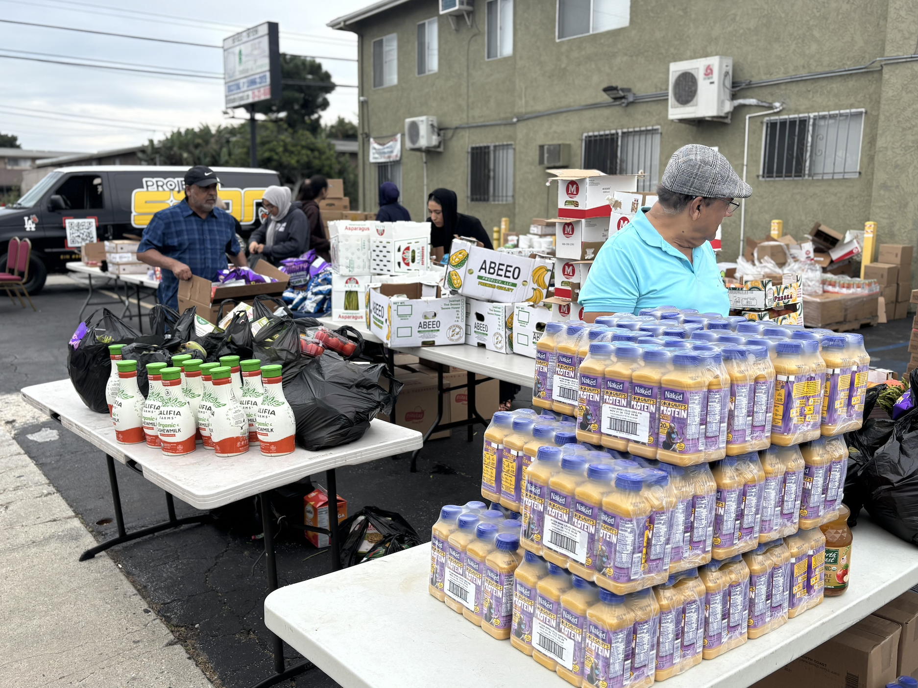People shopping at an outdoor market with tables of bottled drinks, produce, and boxed goods, with a van and a building in the background.