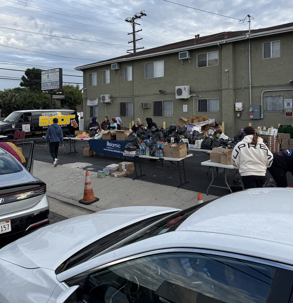 People organizing and distributing supplies at a charitable event outside in a parking lot, with tables holding boxes and bags of goods, a van with 'PROJECT S.C.R' logo, and a green apartment building in the background.