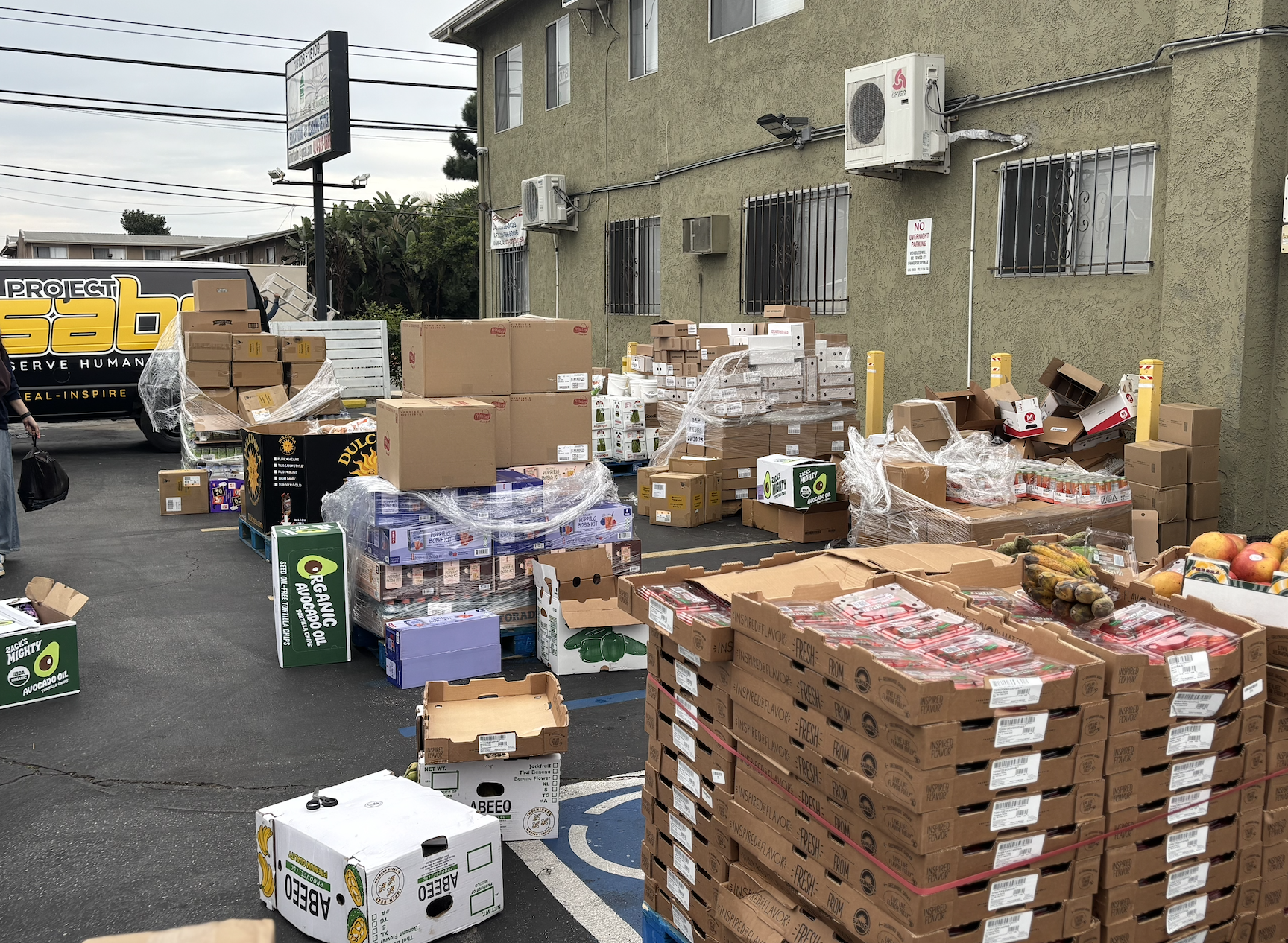 Outdoor scene of a parking lot with numerous stacked boxes and pallets of food and supplies, some wrapped in plastic, near a green building with barred windows and air conditioning units.