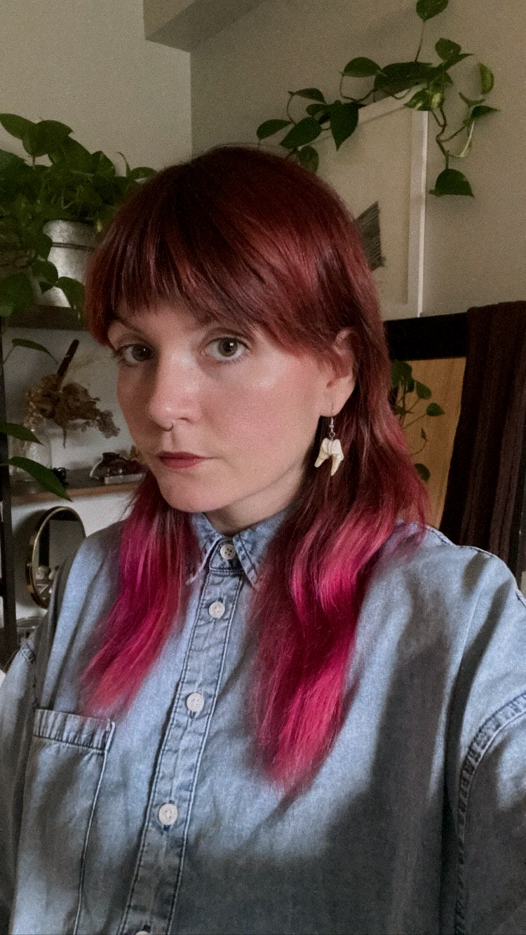 A nonbinary person with red to pink ombre hair, wearing a denim shirt and earrings, standing indoors with potted plants in the background.