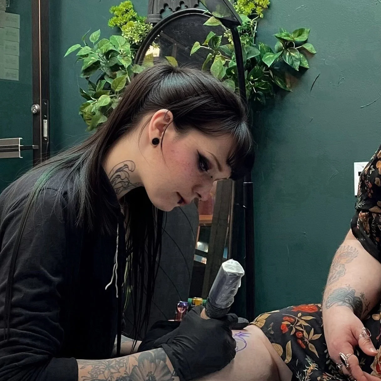 A woman with black hair and tattoos on her neck and arms is tattooing a design on someone's thigh in a tattoo studio, with green plants in the background.