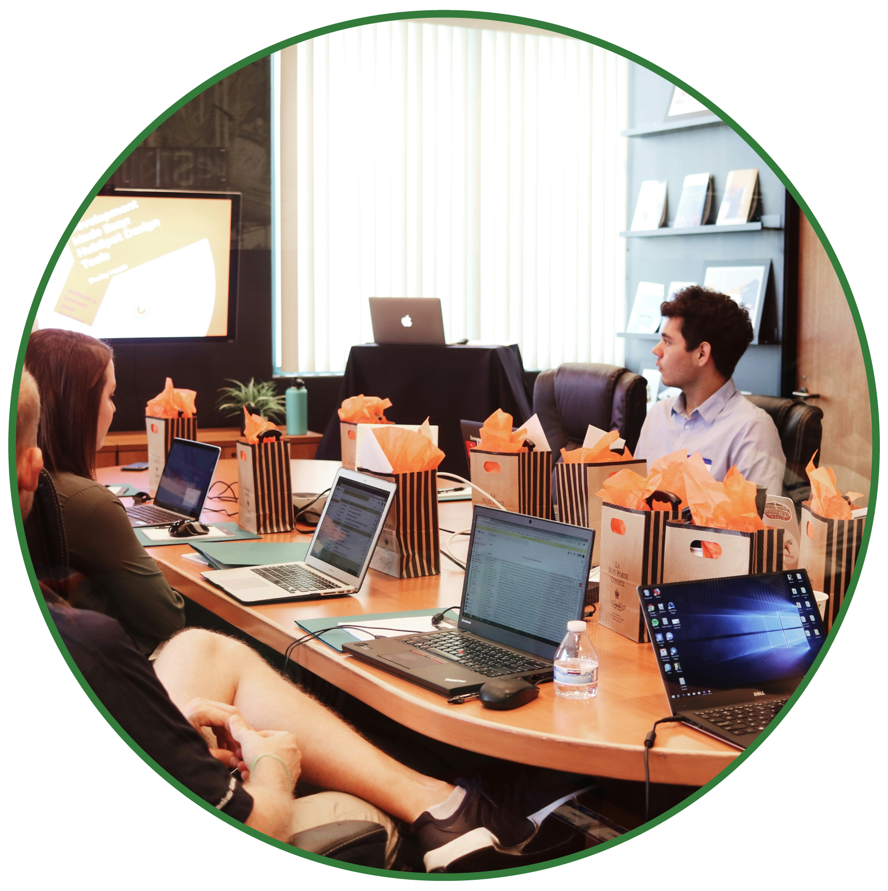 Business meeting in a conference room with people sitting at a large table, using laptops and electronic devices, with gift bags on the table and presentation screen on the wall.