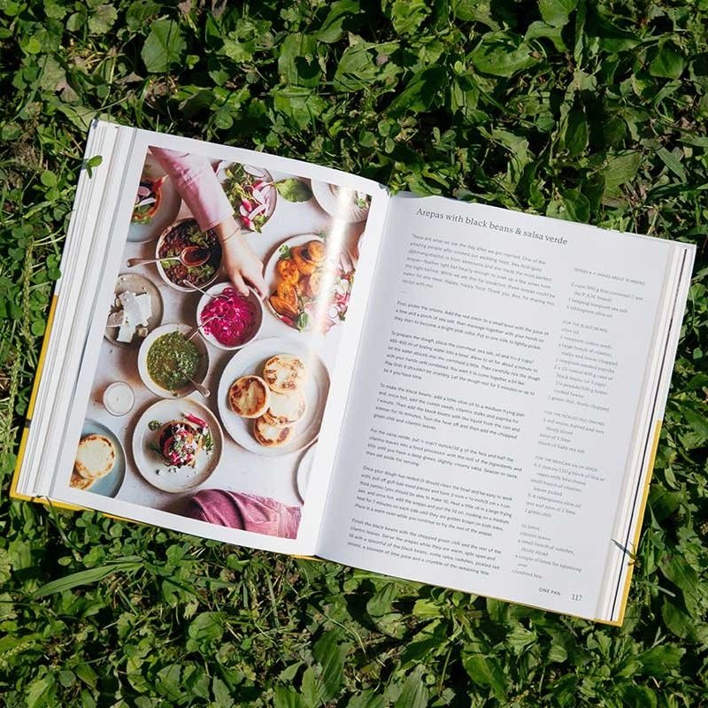 The book One Pot, Pan, Planet book lies open to a bean and salsa verde recipe spread while laying on brightly lit grass.