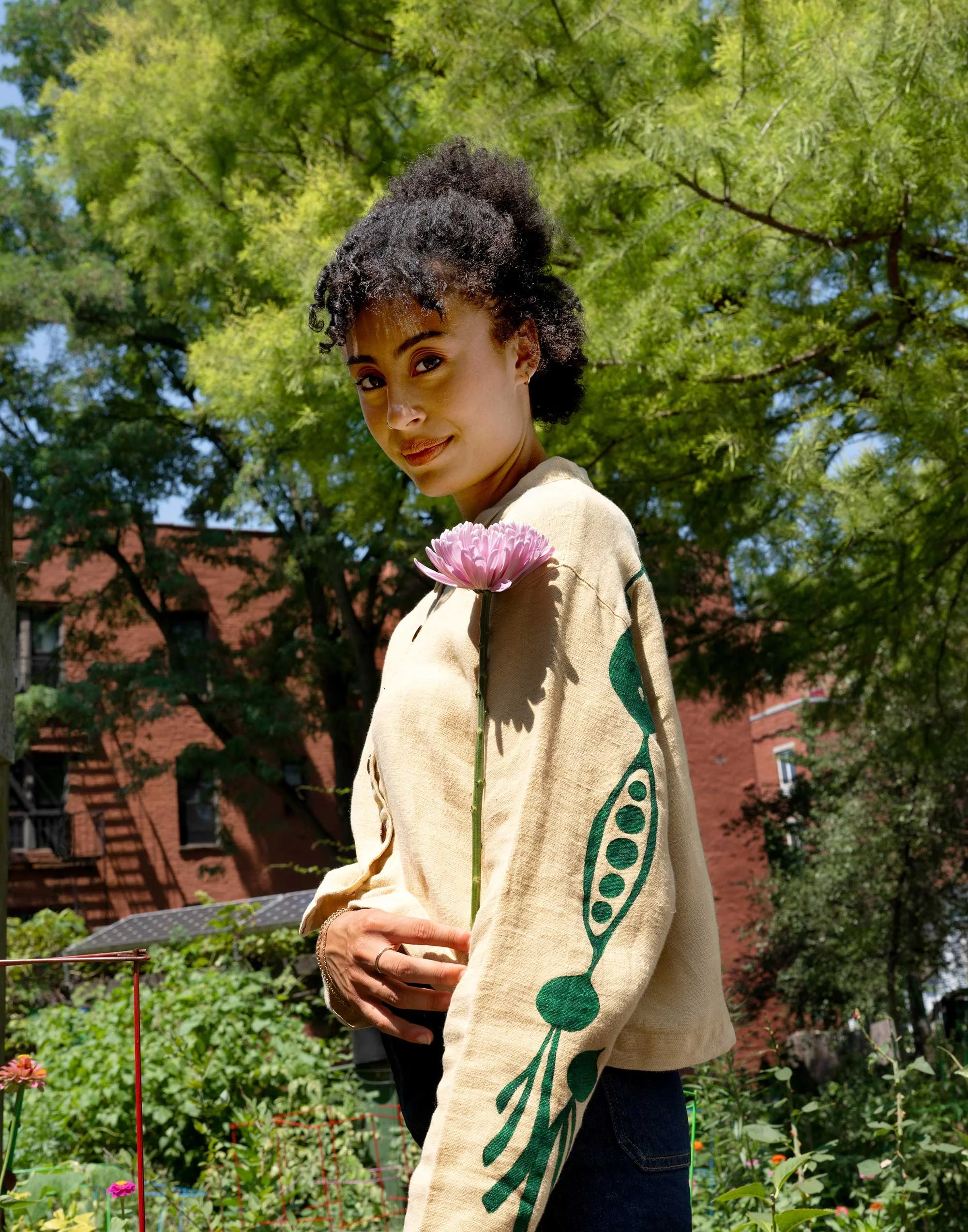 A woman with light brown skin and natural hair stands in a garden holding a pink flower and wearing a light yellow blouse with a veggie motif painted down the back of her arms.