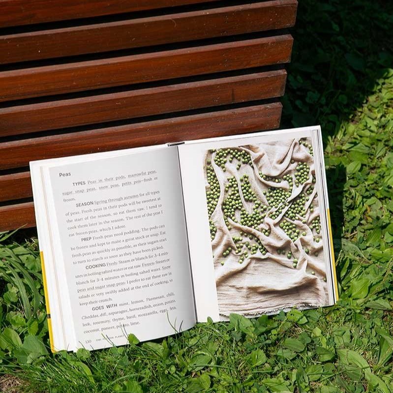 The book One Pot, Pan, Planet book lies open to a spread about peas propped up against a wood slat wall on the grass.