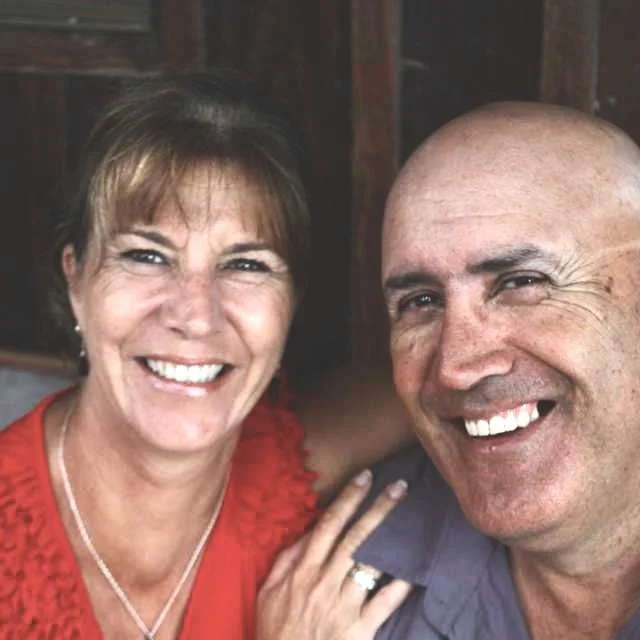 A smiling woman with short brown hair and a man with a shaved head, both smiling warmly, taking a selfie together indoors.