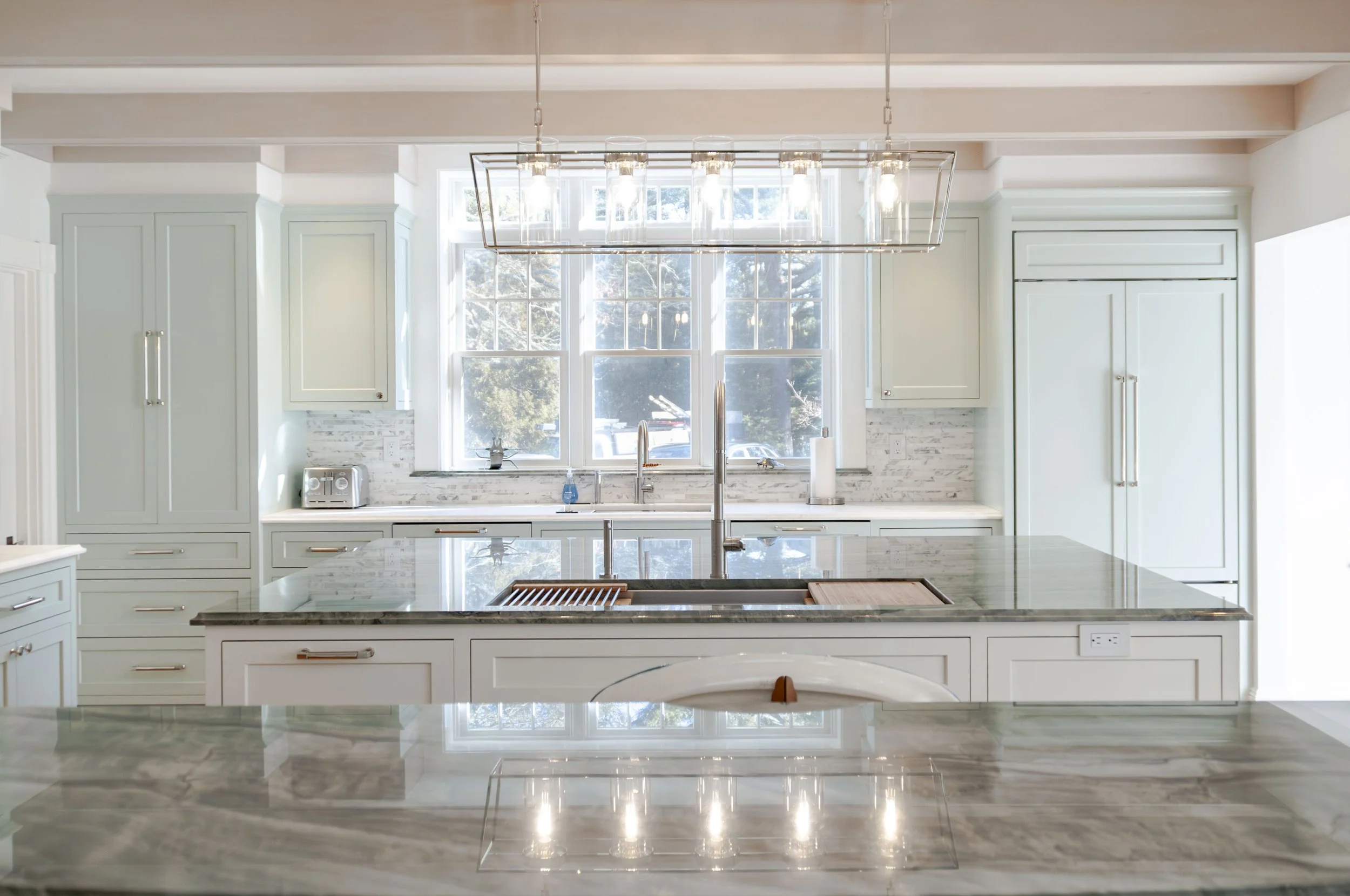 Cape Cod kitchen with marble island, soft green cabinetry, bright window views, and polished pendant lighting.