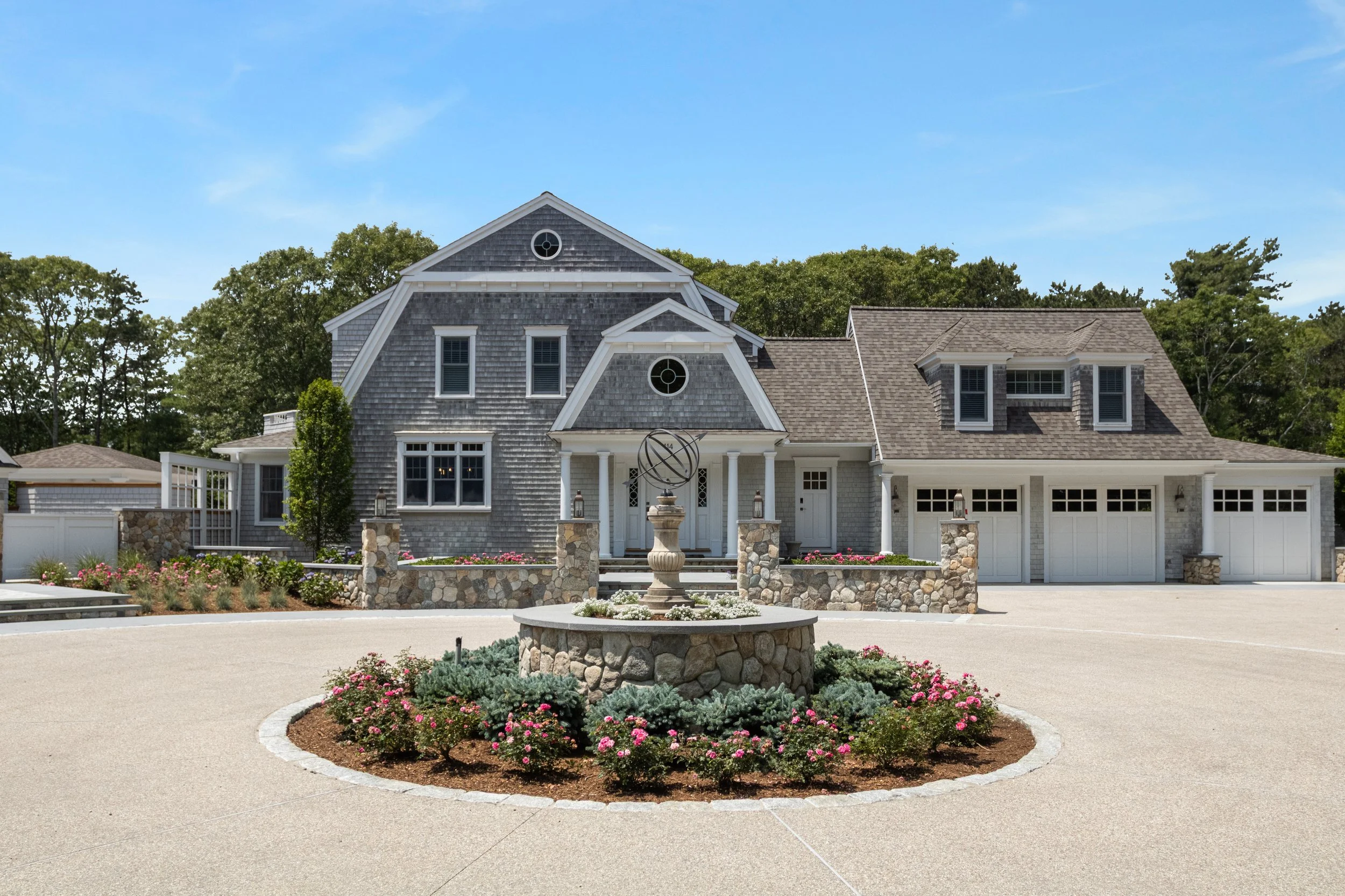 Landscaped circular driveway with stonework and a fountain before a shingle-style Cape Cod home.