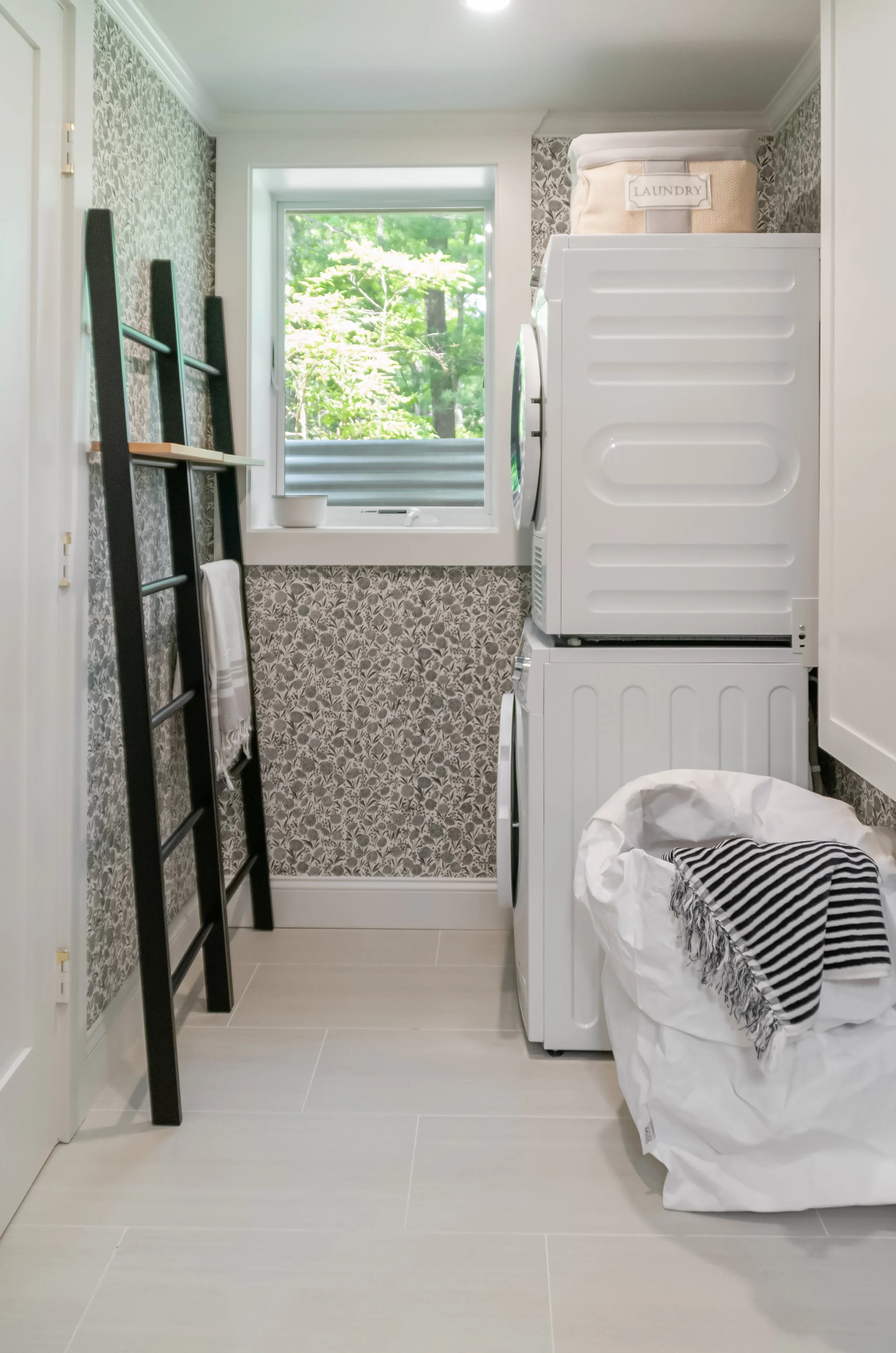 Laundry room with patterned wallpaper, stacked washer and dryer, black towel ladder, and a tree-facing window.