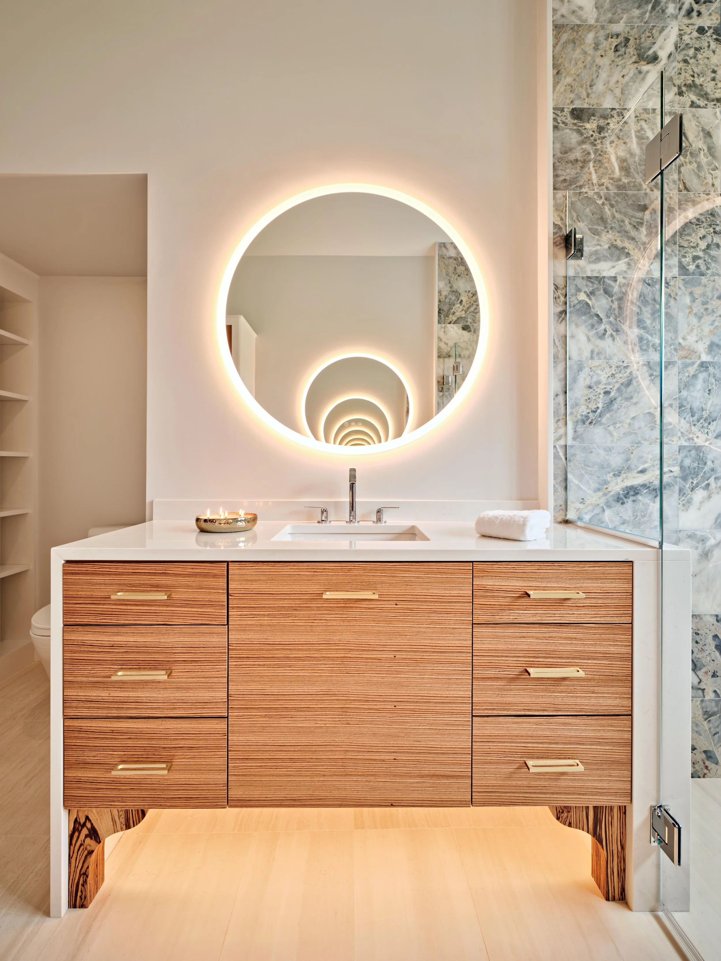 This high-end Cape Cod renovation features an illuminated round mirror, wood vanity, marble shower, and polished bath finishes.