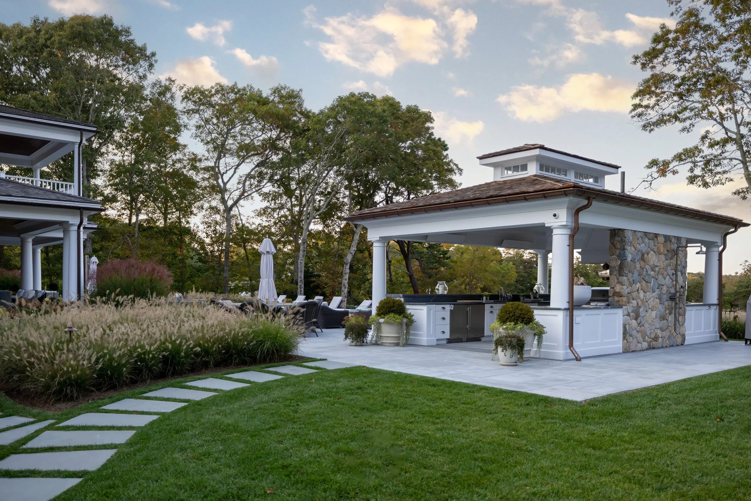 Covered outdoor kitchen at a custom coastal home with stone accents, white columns, and Cape Cod landscaping.