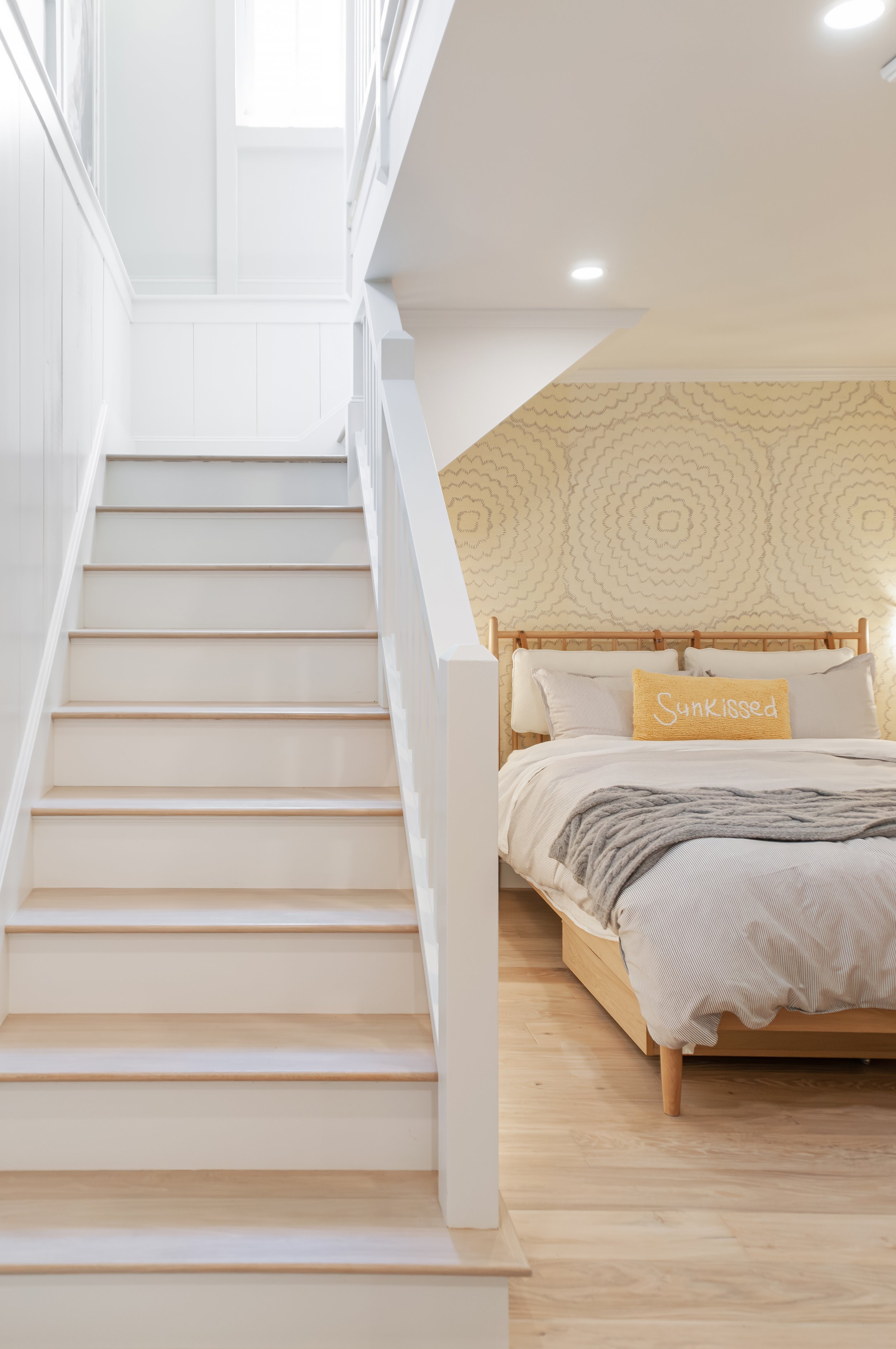 White staircase beside a cozy bedroom with wood floors, patterned wallpaper, and a sunlit skylight.