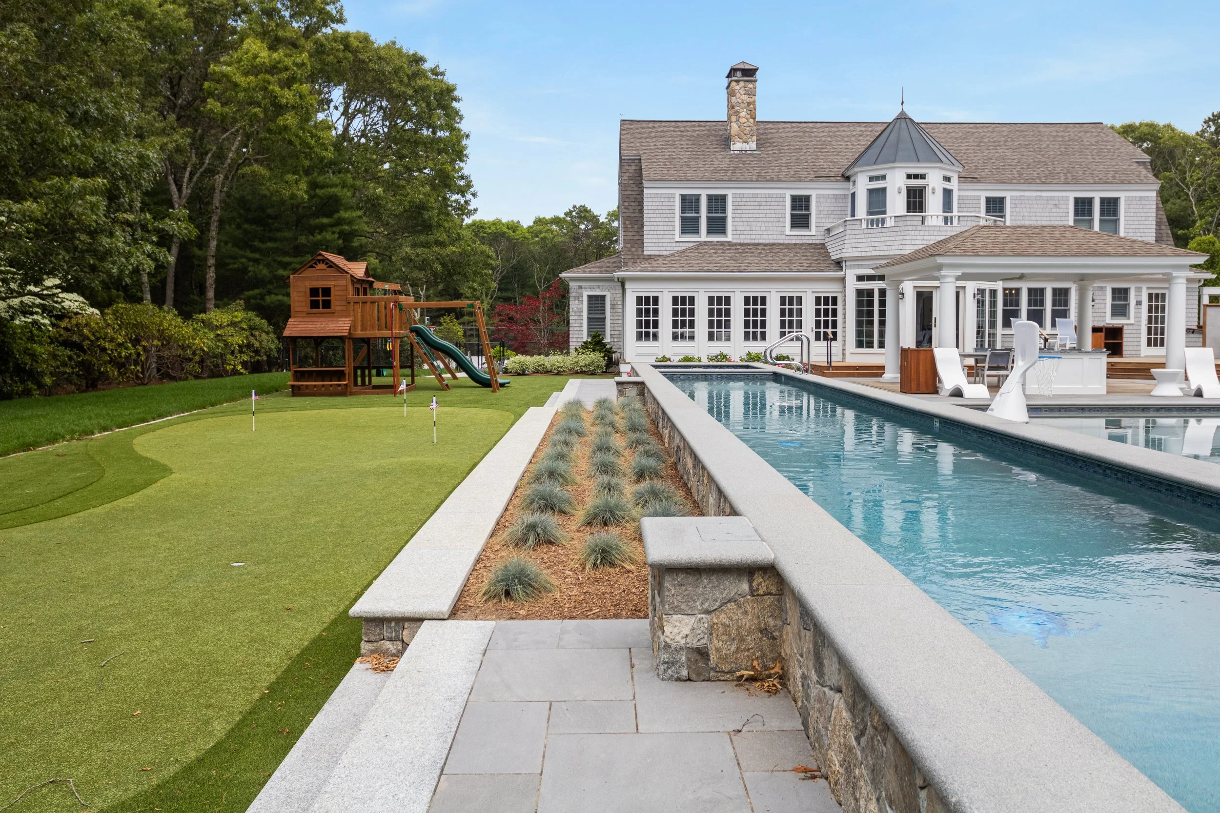 Expansive outdoor living area featuring a custom lap pool, putting green, stone retaining walls, and luxury landscaping surrounding this Cape Cod custom home.