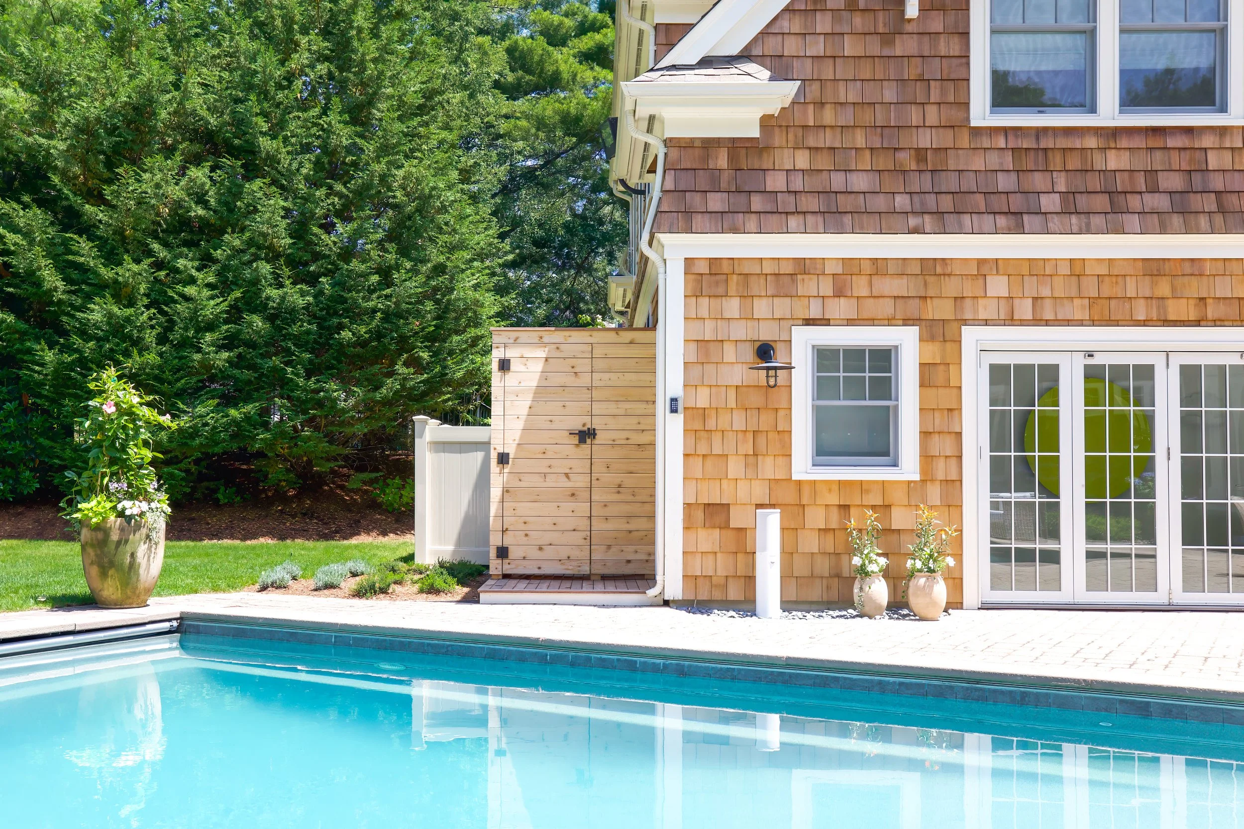 Poolside view of a shingled structure, French doors, cedar gate, and Cape Cod pool house design.