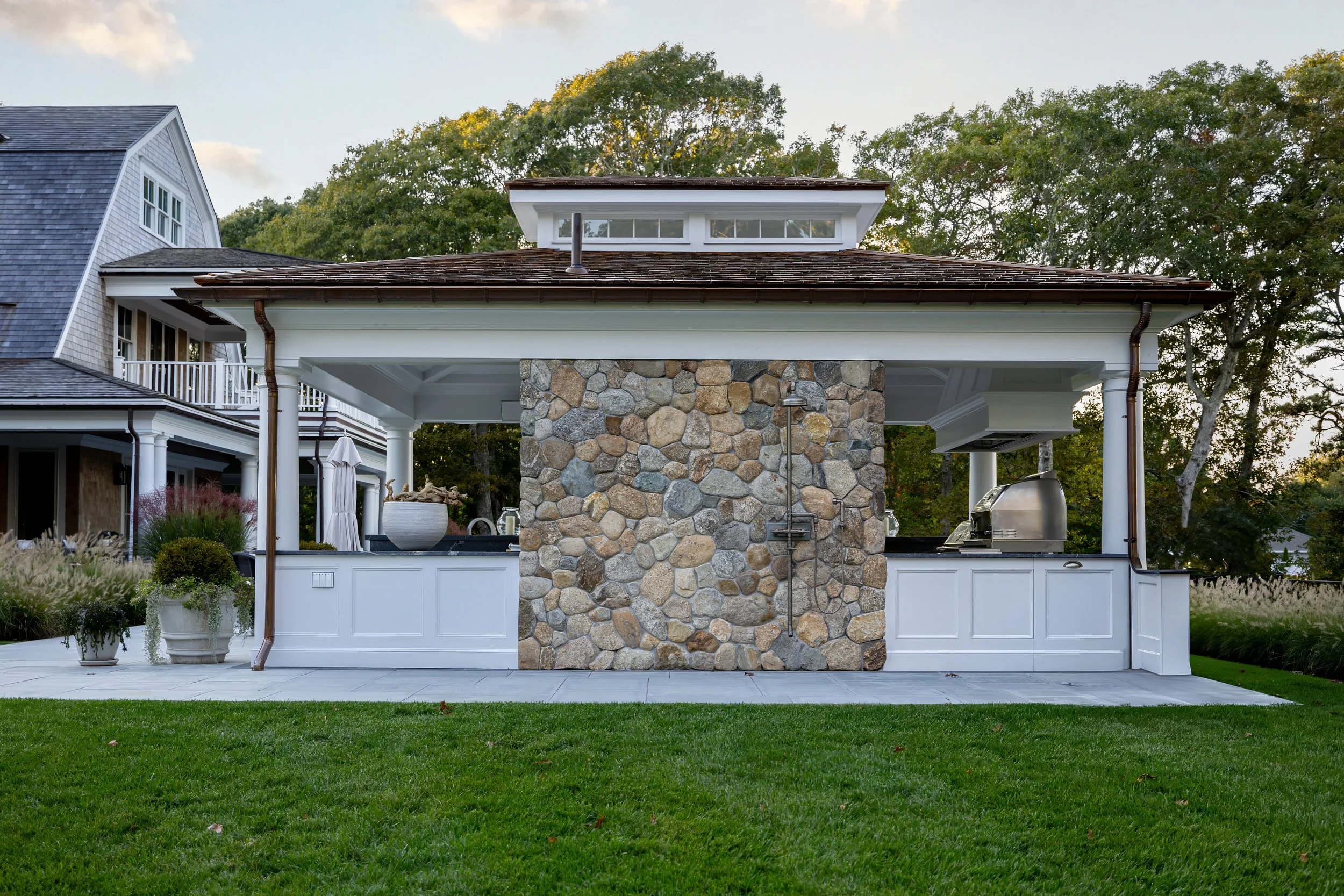 Covered outdoor kitchen with stone shower wall, white cabinetry, copper gutters, and Cape Cod lawn.