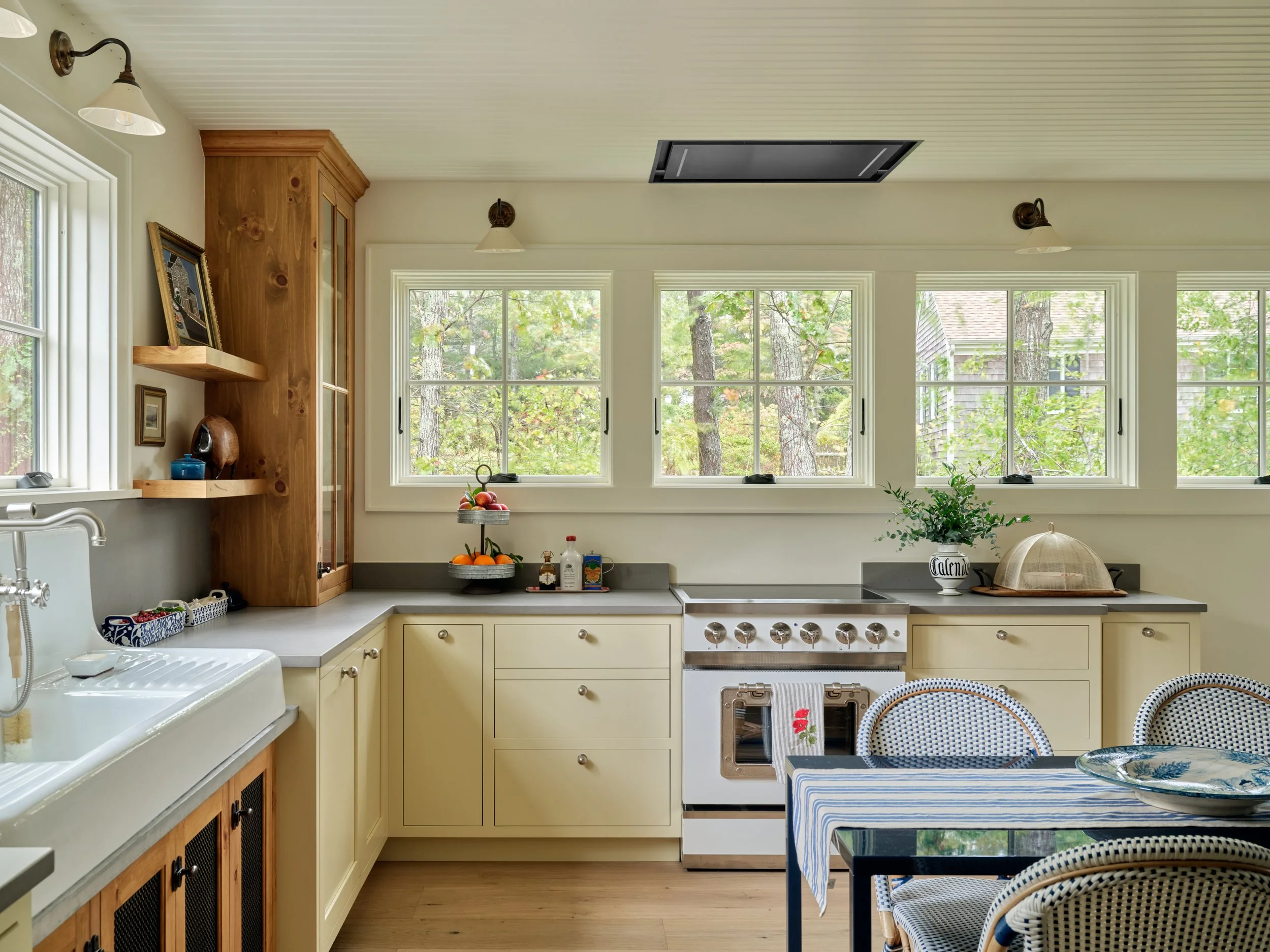 Framed by oversized windows, this Sea View Builders kitchen features custom cabinetry, a farmhouse sink, wood shelving, and timeless Cape Cod design.