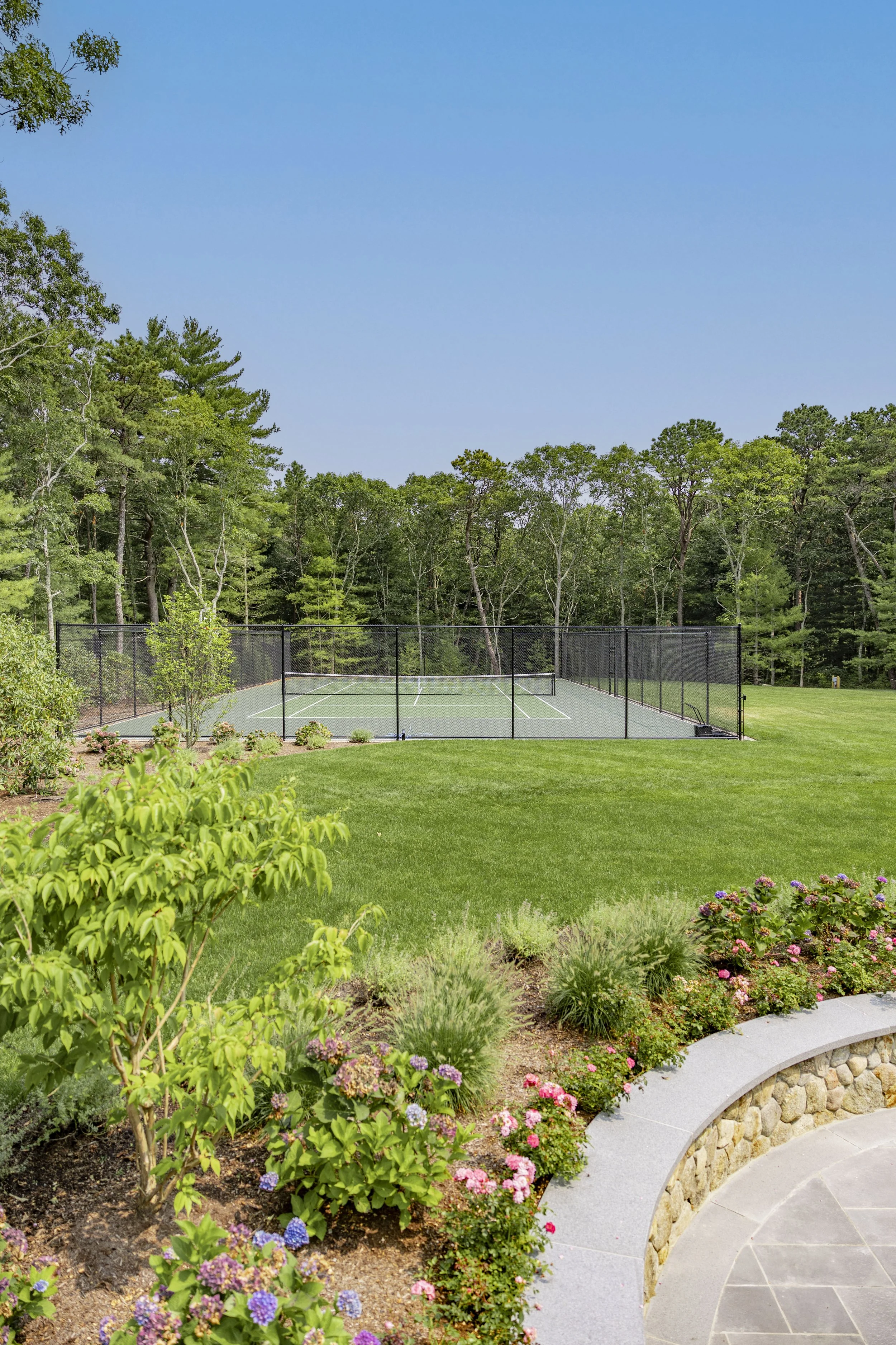 Landscaped Cape Cod property with tennis and pickleball courts, manicured lawn, gardens, and wooded backdrop.