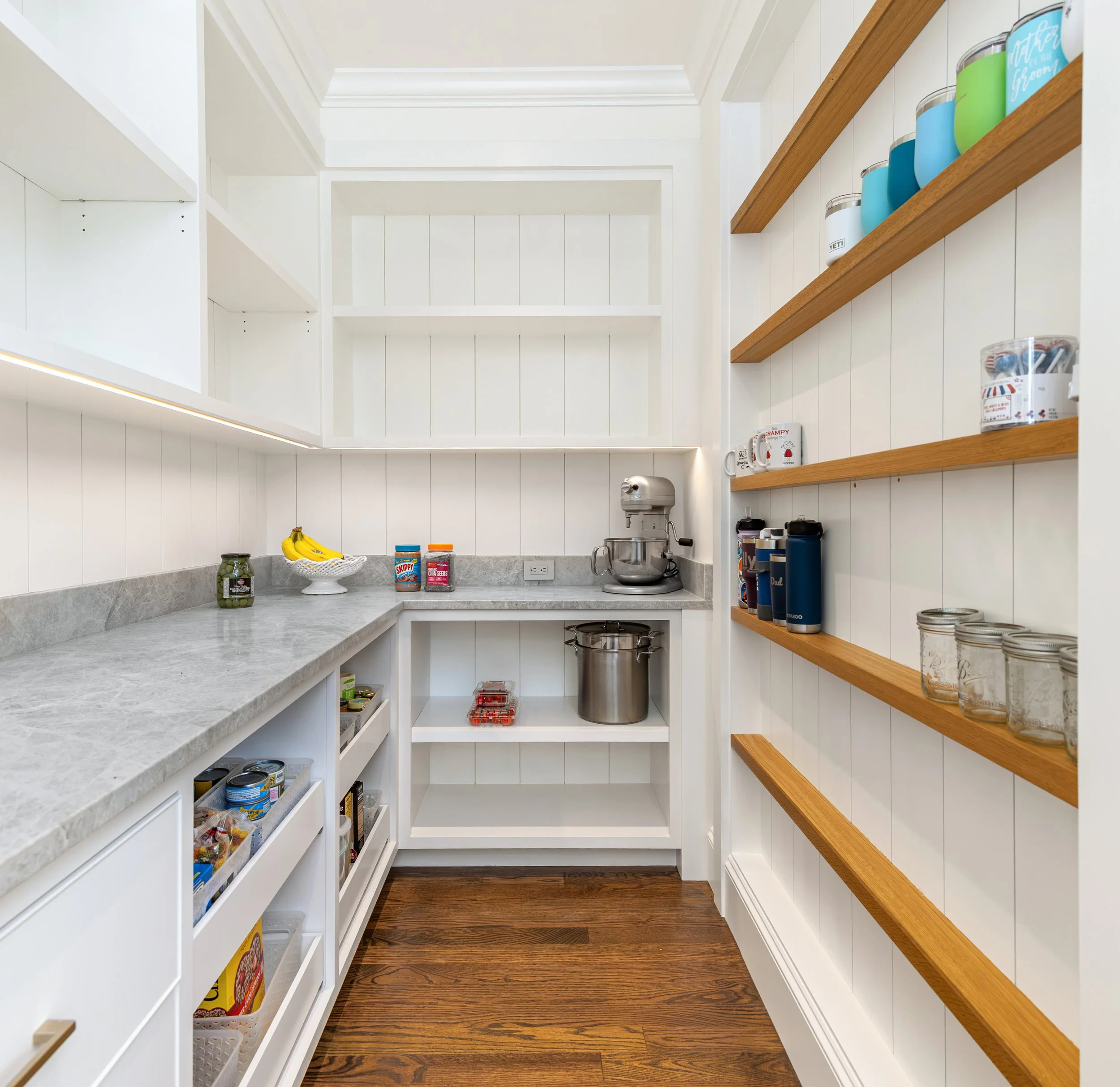 Custom walk-in pantry with marble countertops, open wood shelving, built-in storage, and shiplap walls for organized kitchen essentials.