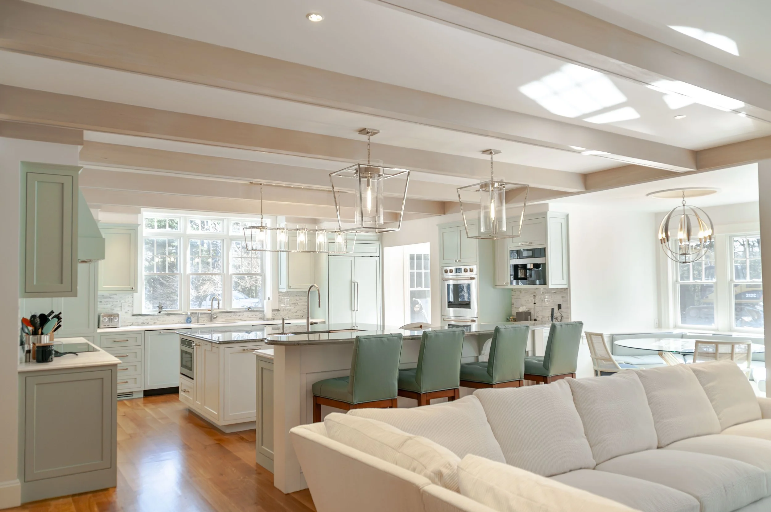 Open Cape Cod living room with white sectional, blue patterned rug, beamed ceiling, and kitchen beyond.
