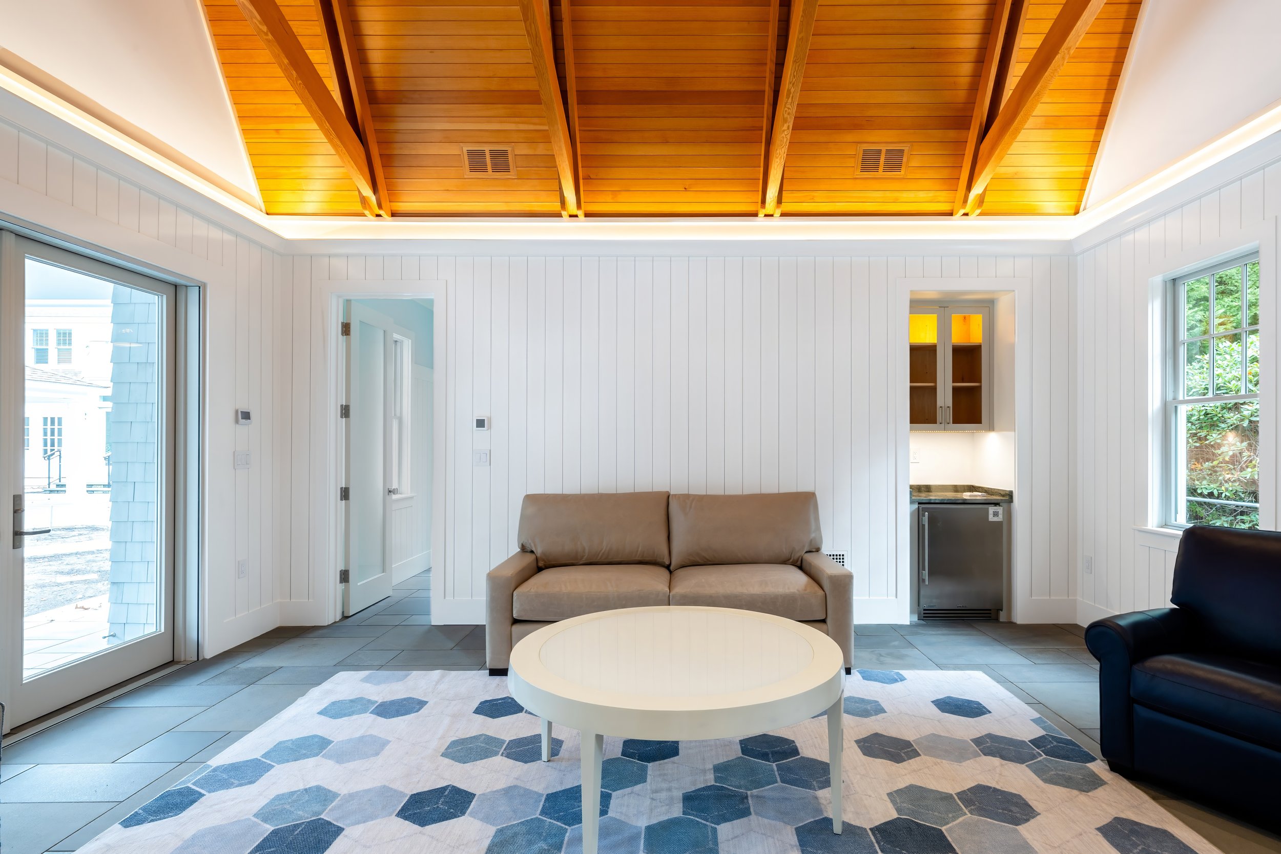 Cape Cod pool house lounge with illuminated wood celing, white paneling, and sliding glass doors