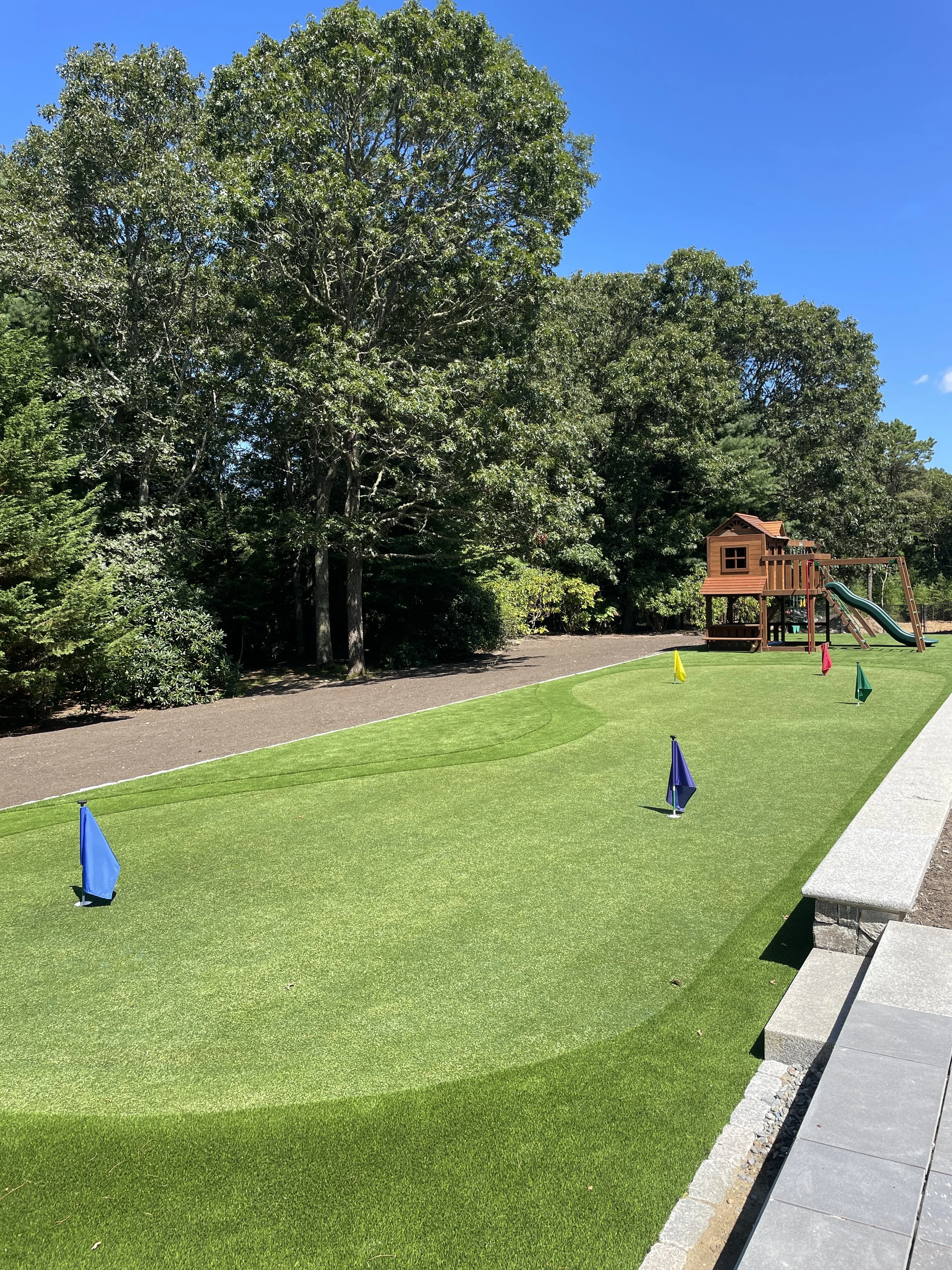 Backyard putting green with golf pins, wooded border, and playset at a Cape Cod property.
