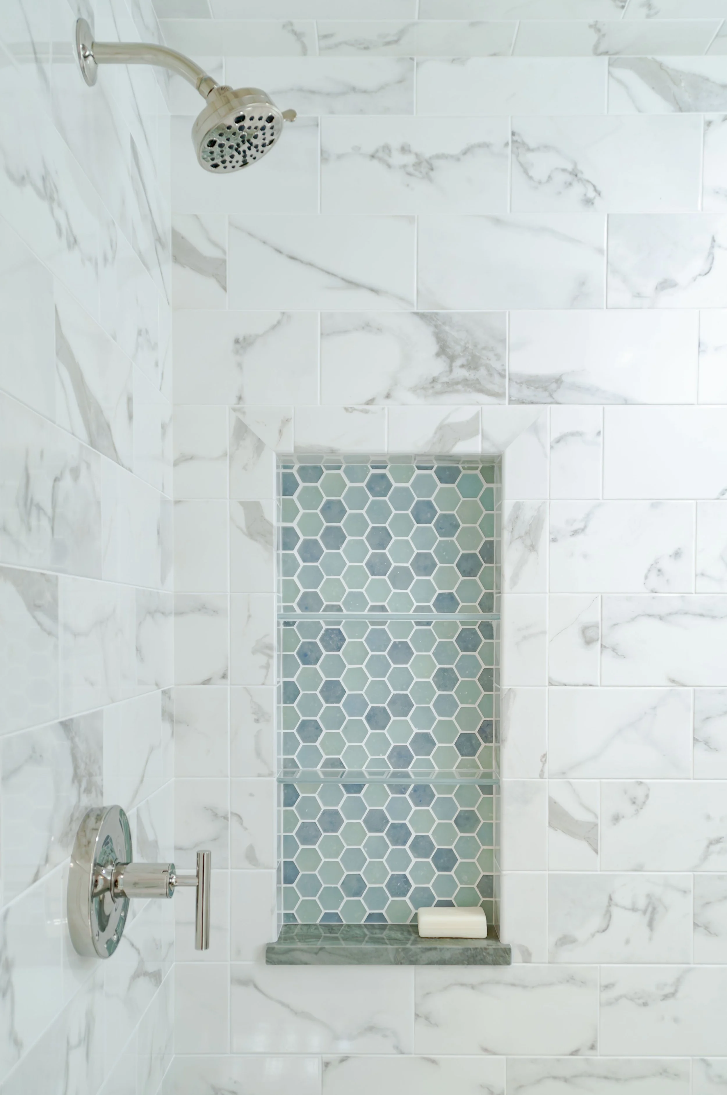 Cape Cod bathroom shower with marble-look tile, blue hexagon niche, polished fixtures, and coastal custom home detail.