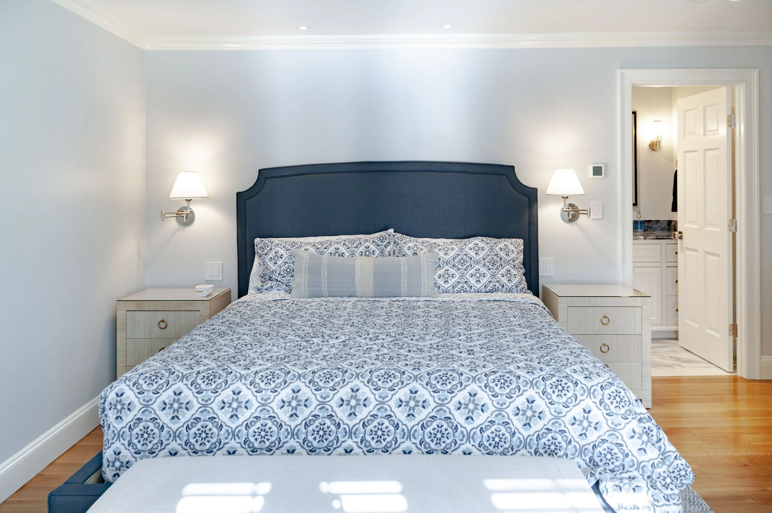 Luxury Cape Cod bedroom with blue upholstered headboard, patterned bedding, wall sconces, and adjoining bathroom.