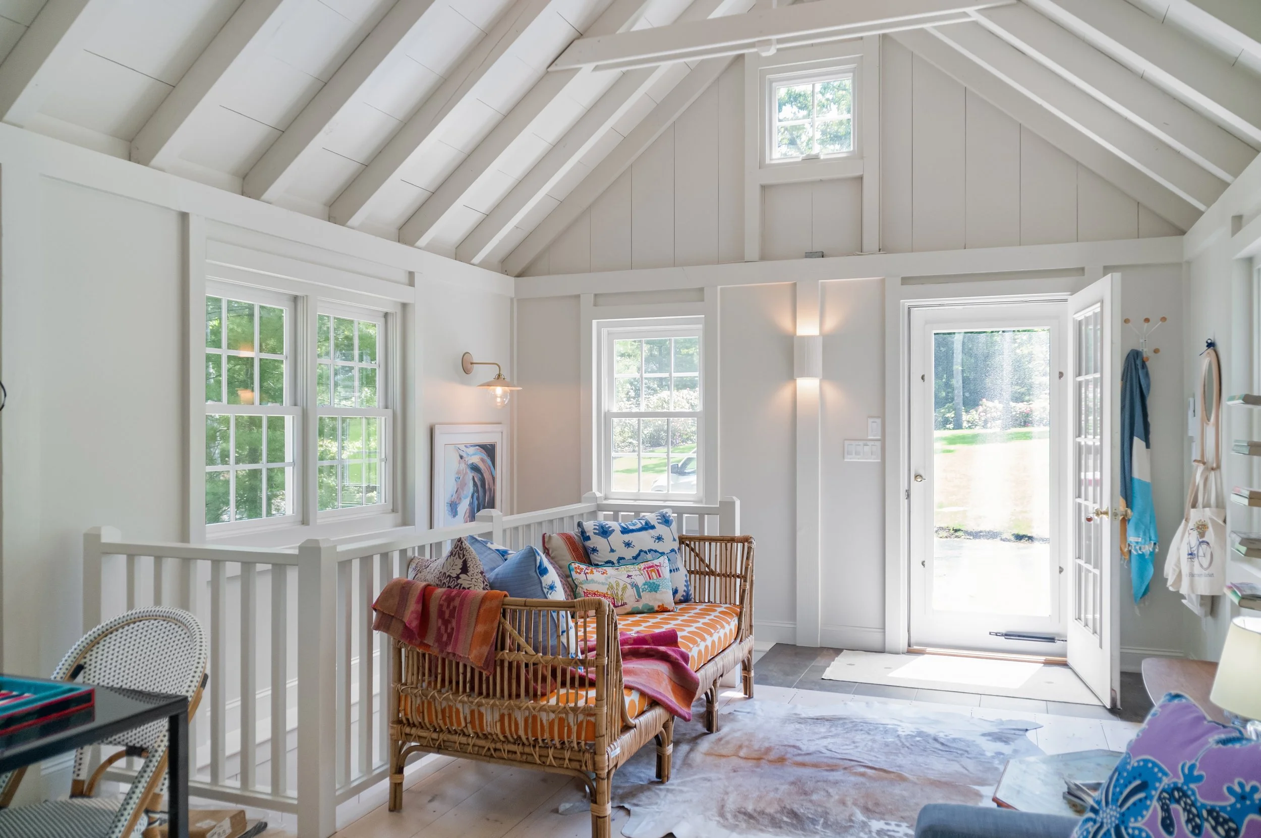 Vaulted sitting area with white paneled walls, exposed rafters, colorful wicker seating, and an open glass door.
