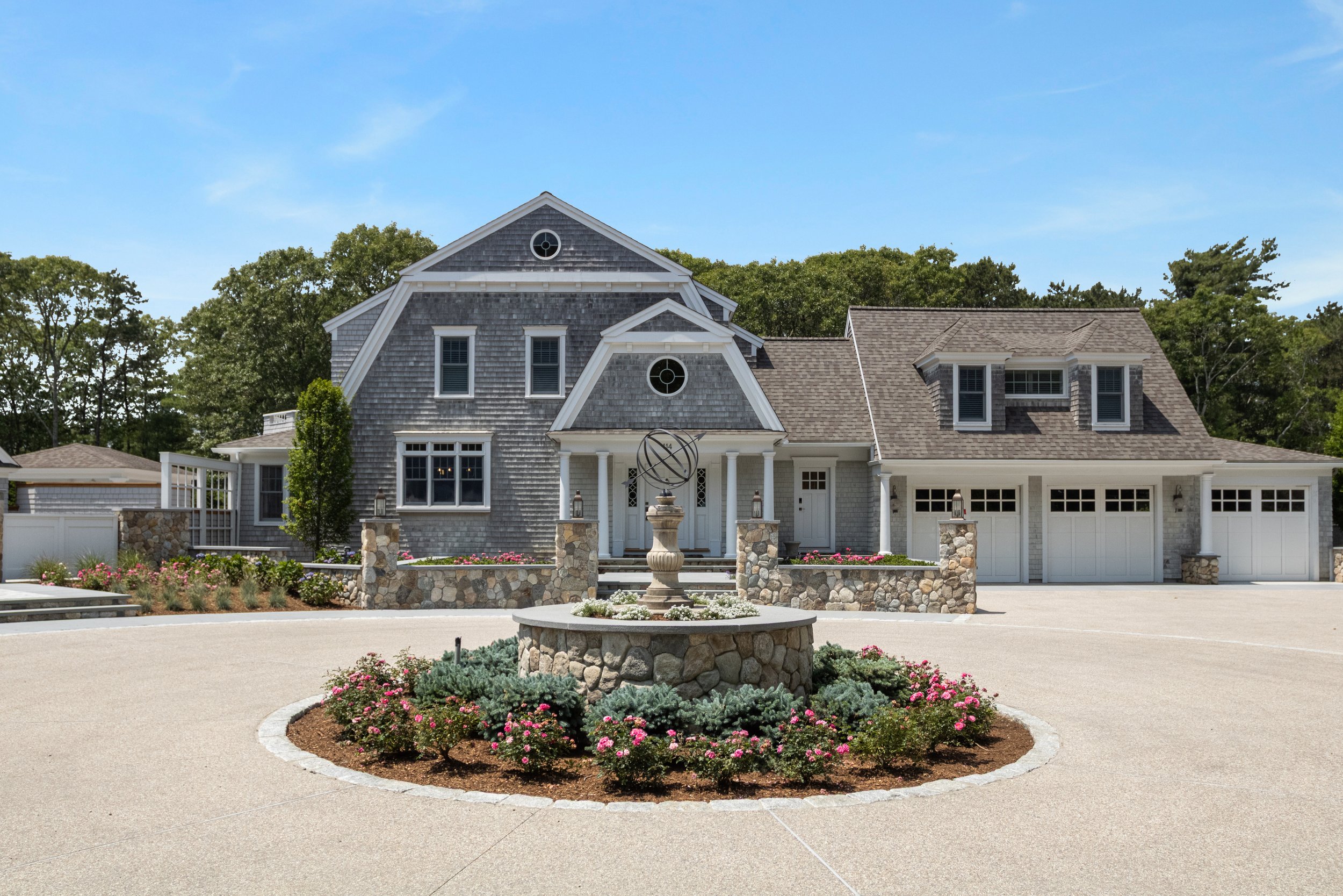 Welcoming front elevation showcases a custom Cape Cod home with cedar shingles, stone walls, elegant landscaping, and a circular driveway with a centerpiece fountain.