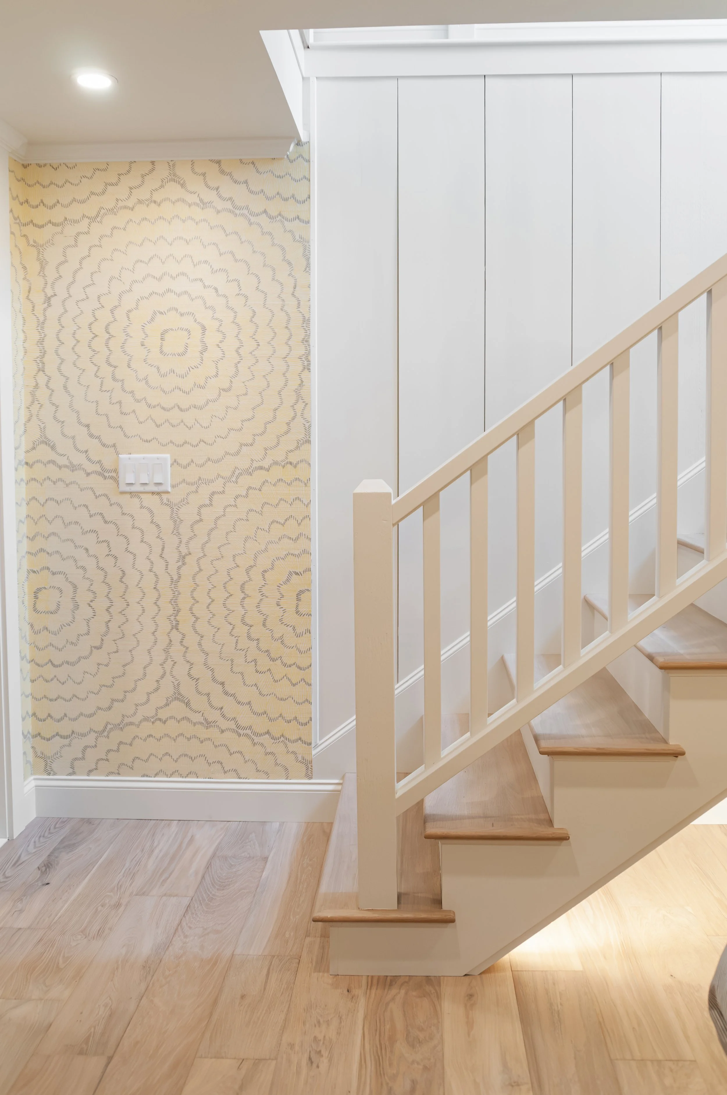Light wood staircase with white railings, vertical paneling, and patterned wallpaper in a bright coastal interior.