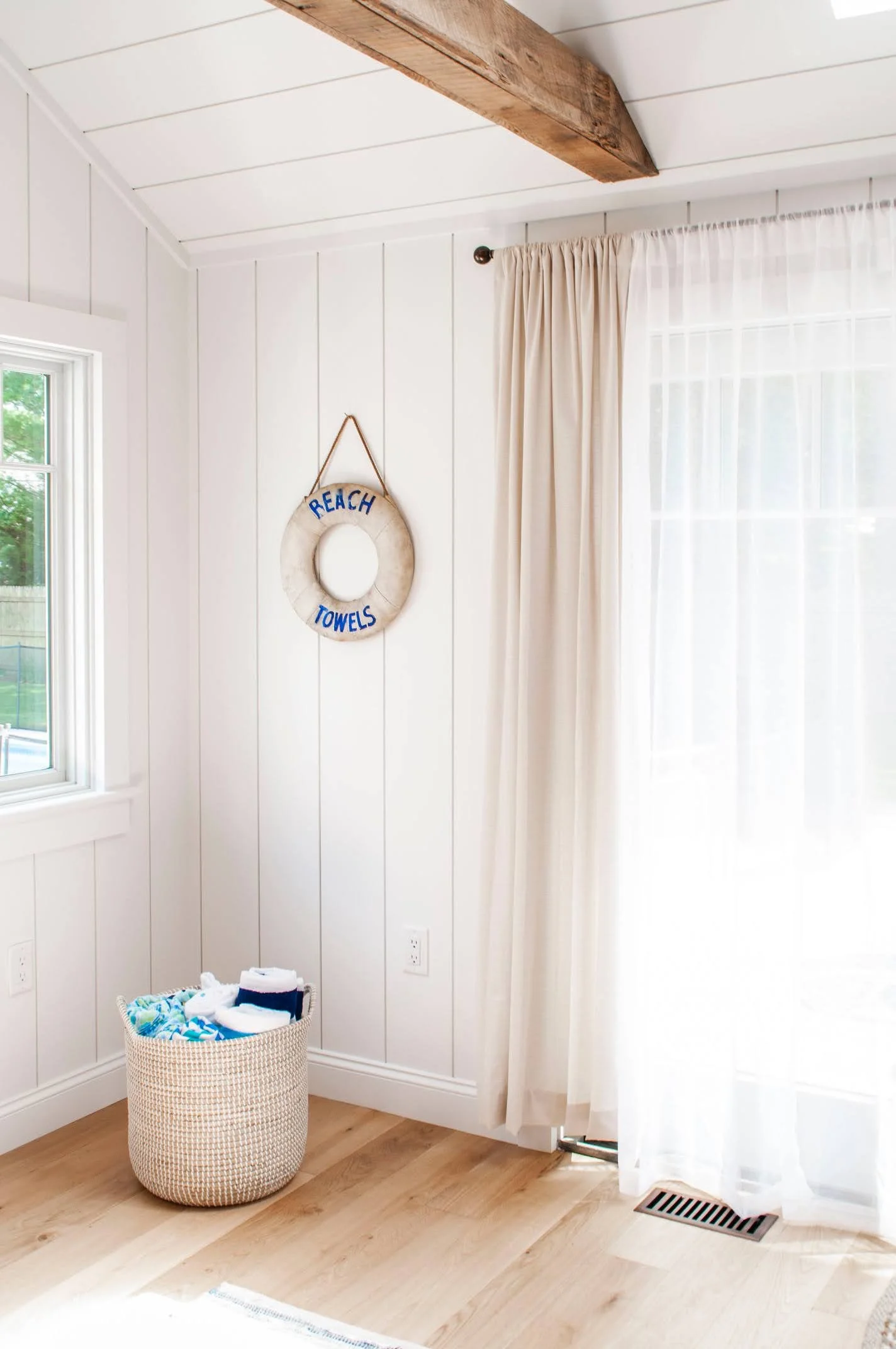 Bright coastal interior with vertical paneled walls, wood ceiling beam, woven basket, and sheer curtains.