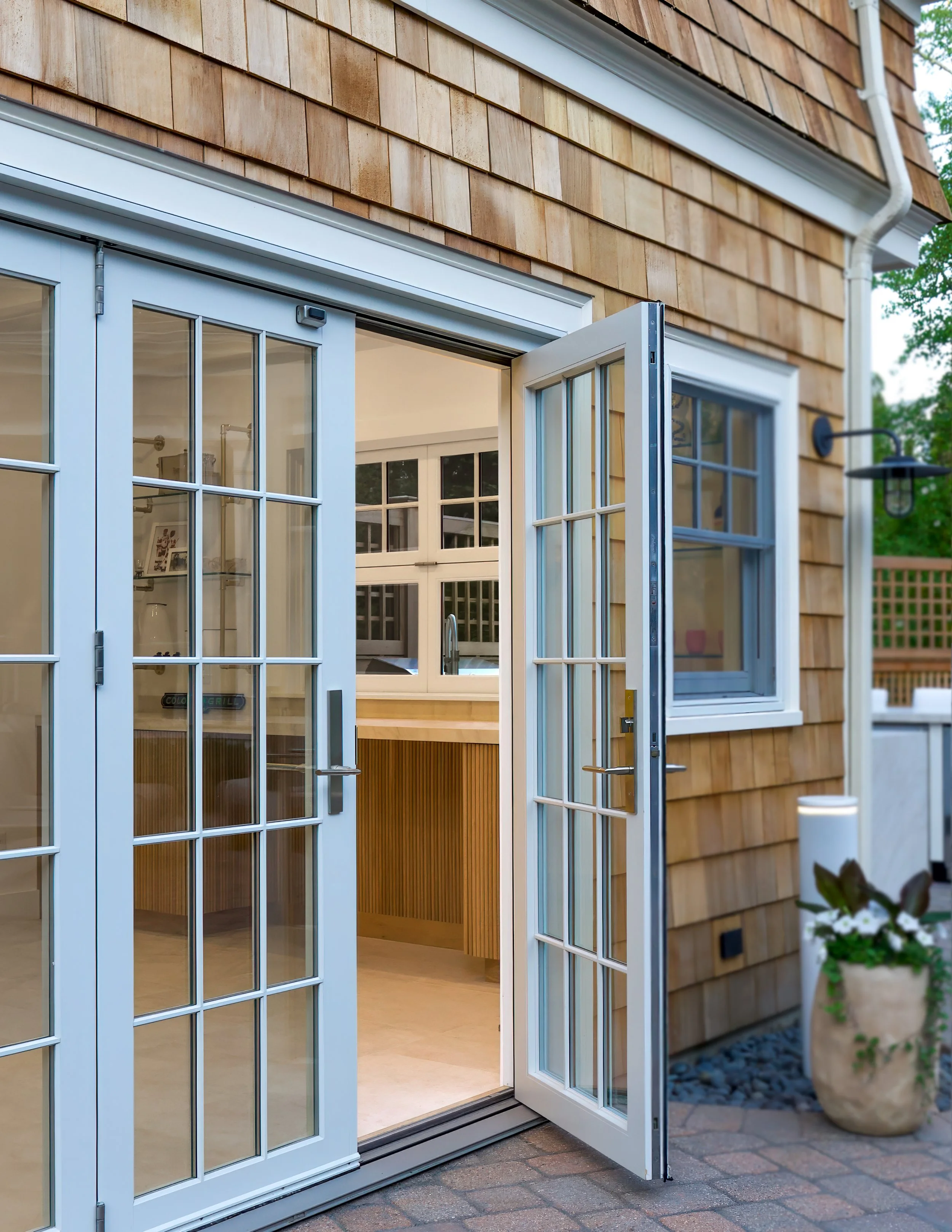 Open French doors reveal a pool house interior beside cedar shingles and a patio entry.