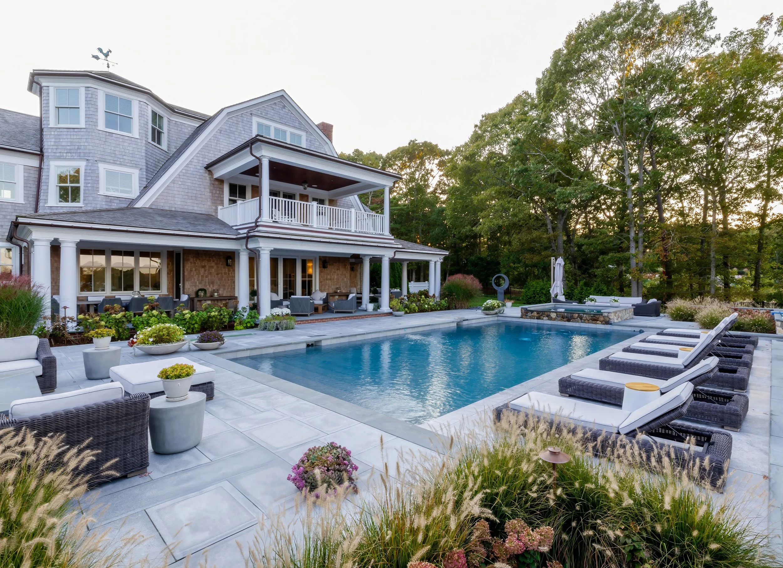 Cape Cod shingle home with poolside lounge chairs, layered plantings, covered porch, and stone patio.