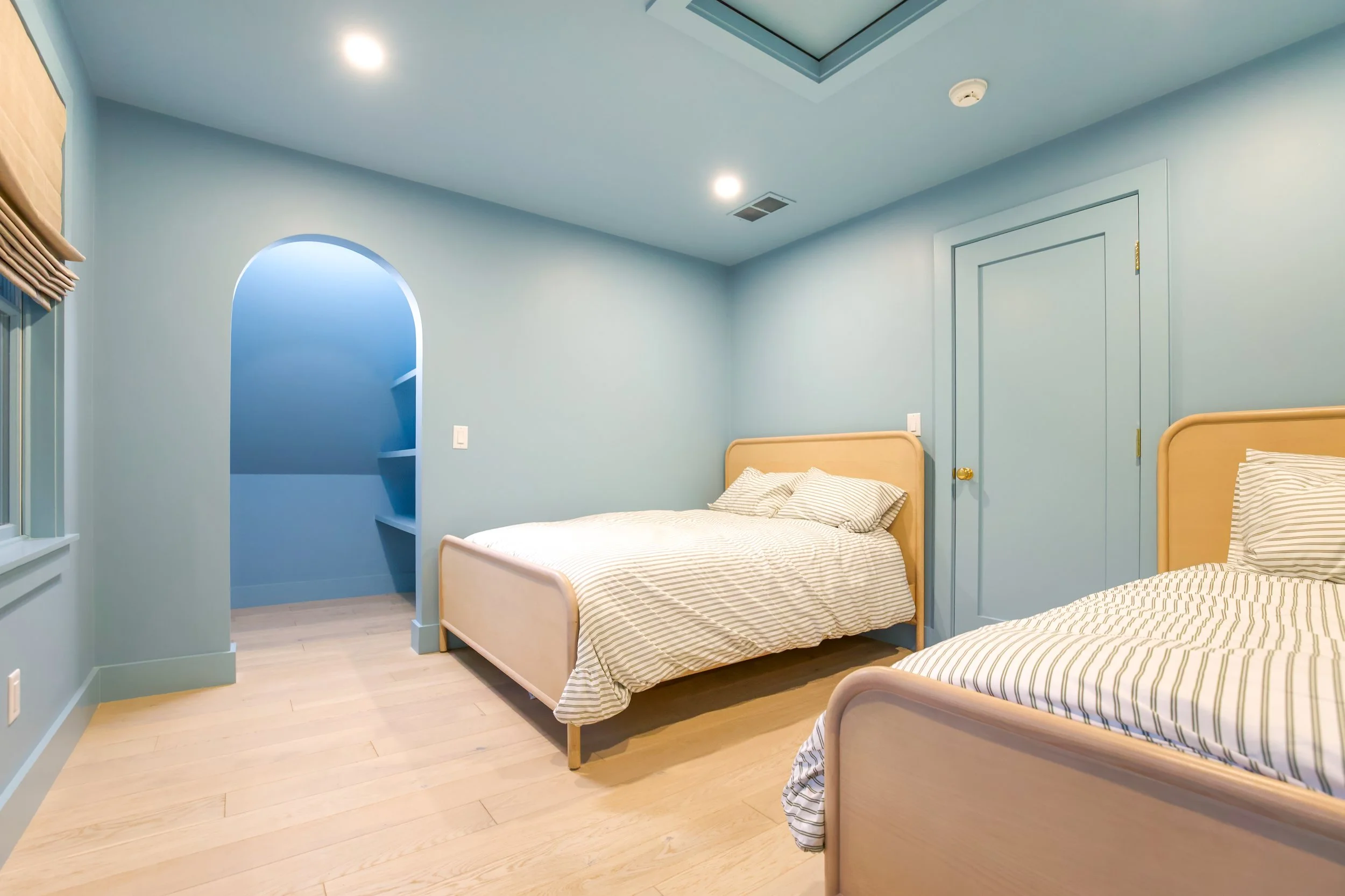 Luxury children's bedroom featuring twin upholstered beds, custom millwork, arched entry, natural wood flooring, and coastal interior design in a Cape Cod custom home.