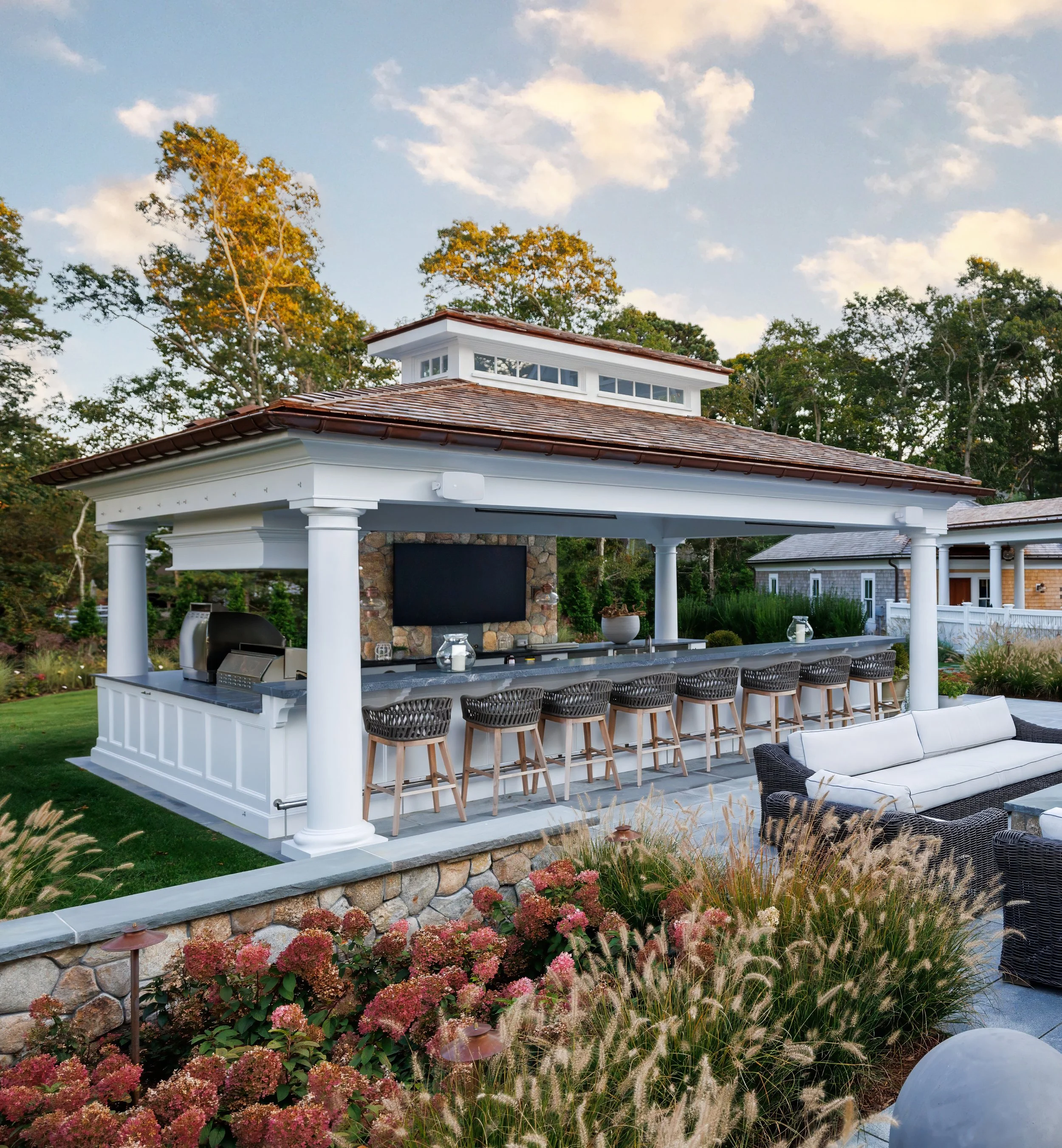 Covered outdoor bar with white columns, long counter seating, stone wall, and hydrangea-lined landscaping.