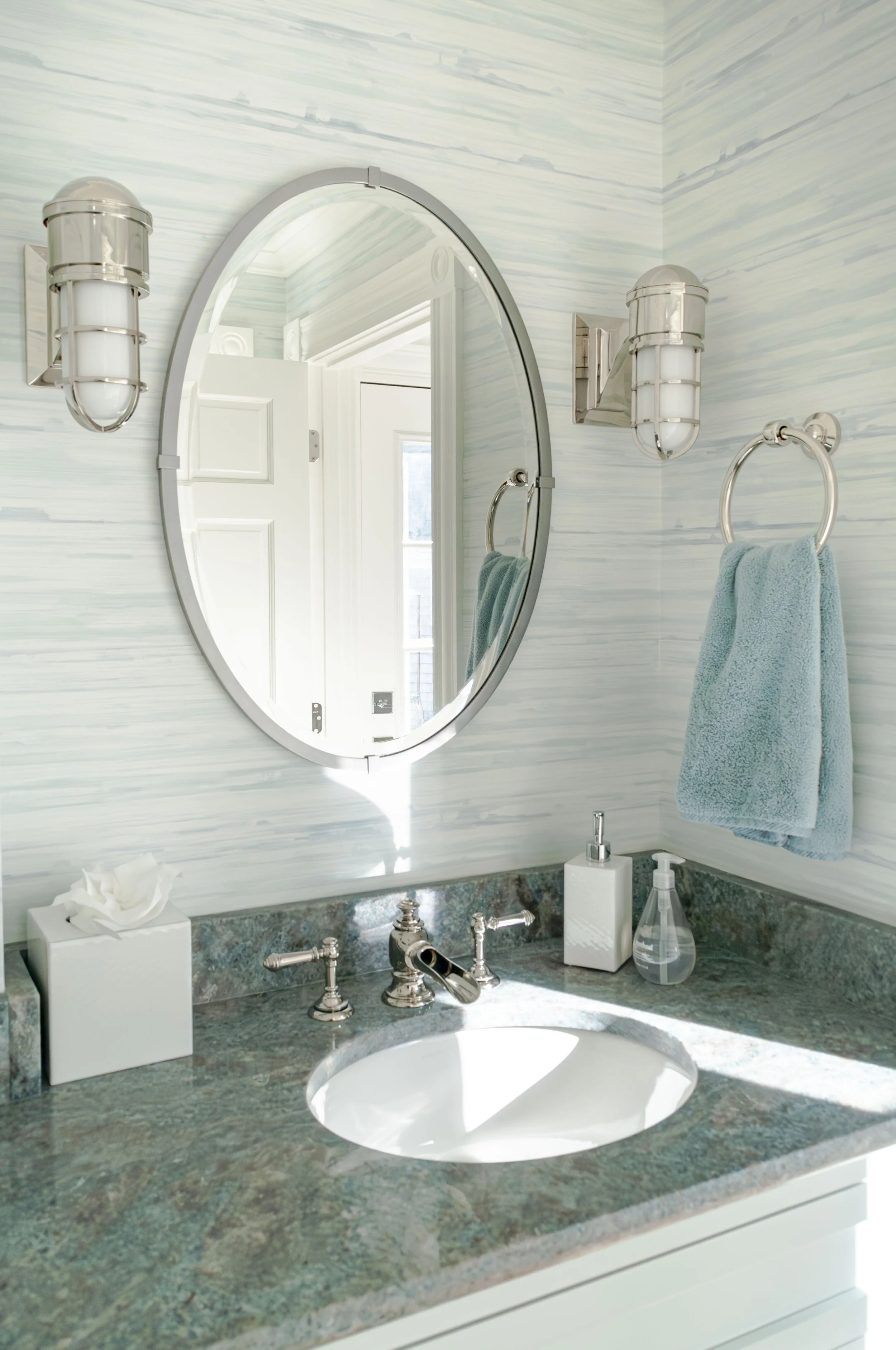 Cape Cod bathroom vanity with oval mirror, blue-green stone counter, wall sconces, and coastal wallpaper.