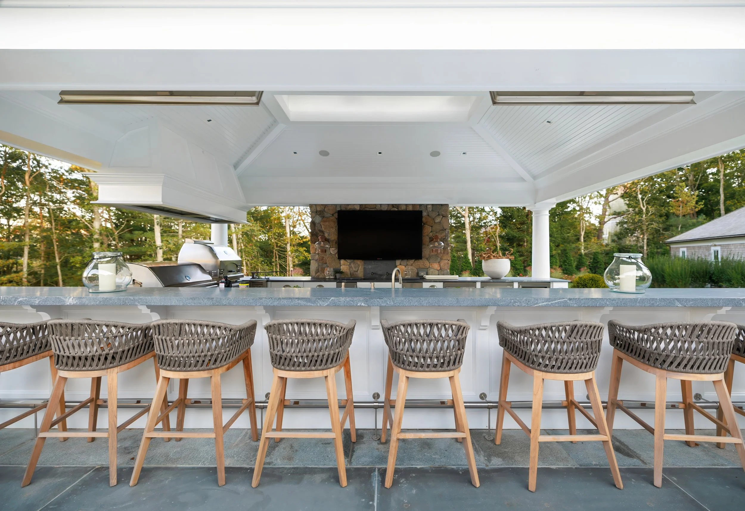 Covered outdoor bar with woven stools, stone fireplace, grilling area, and polished Cape Cod entertaining space.