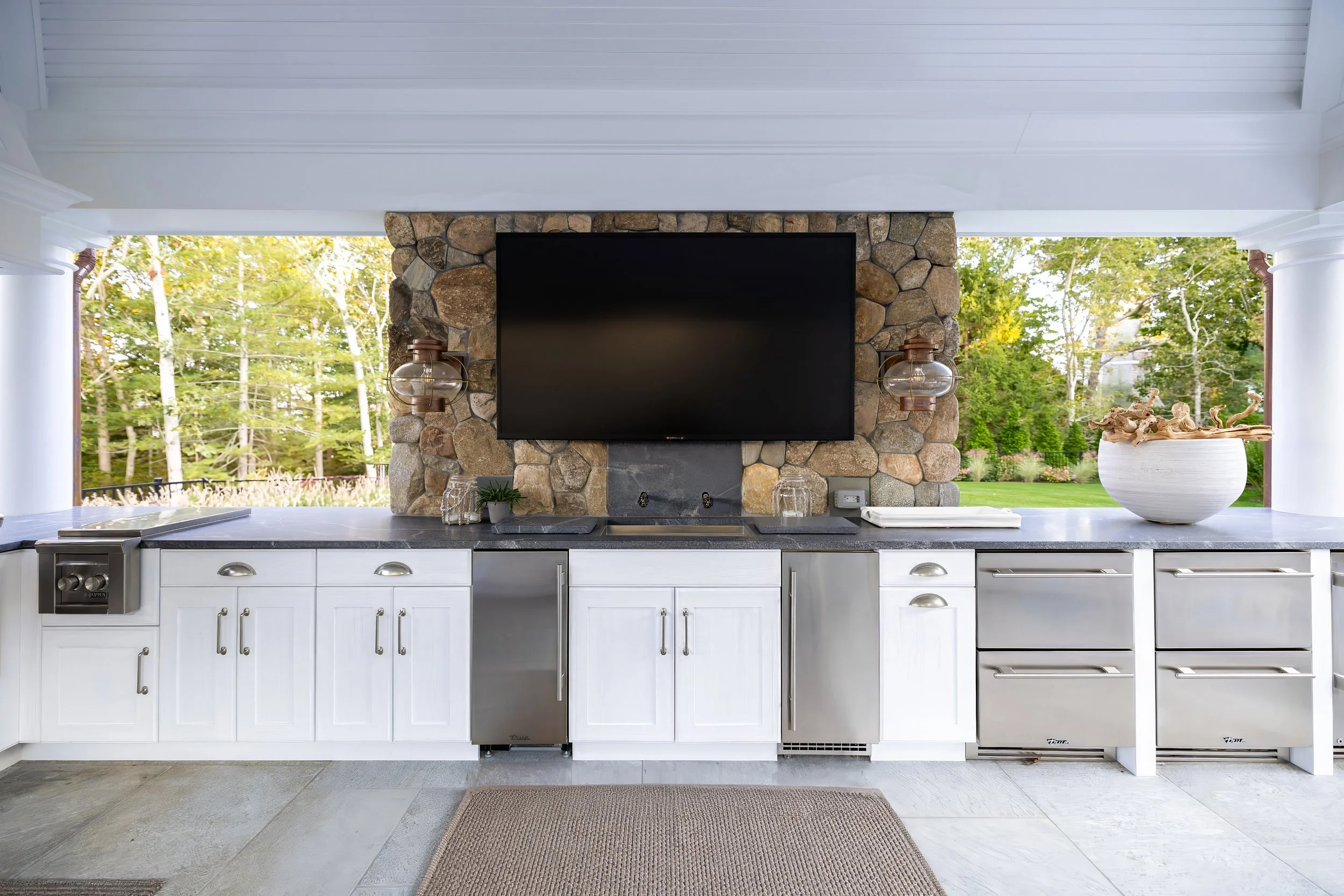 Outdoor kitchen with stone TV wall, white cabinetry, beverage drawers, and wooded Cape Cod views.