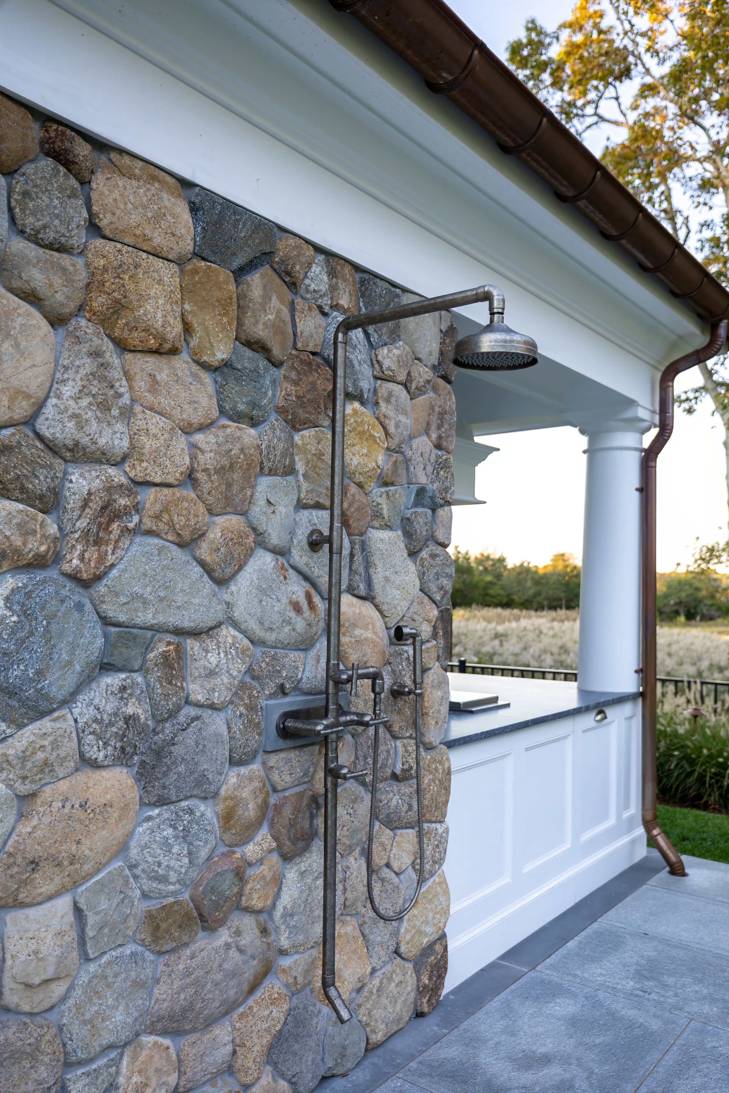 Stone outdoor shower with metal fixtures, white cabinetry, copper gutters, and open Cape Cod views.