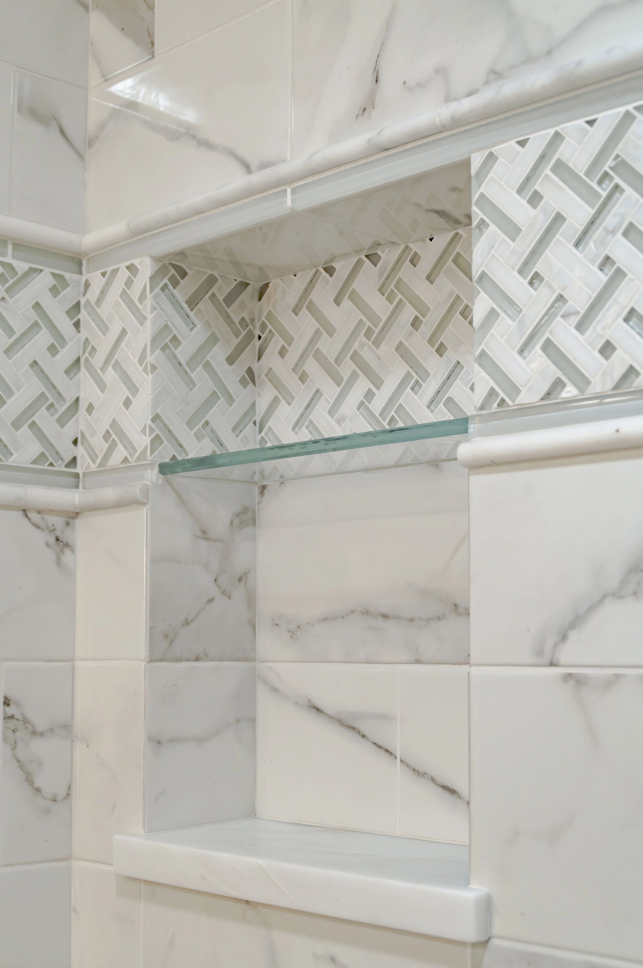 Luxury Cape Cod shower niche with marble-look tile, mosaic accent band, glass shelf, and custom coastal detailing.