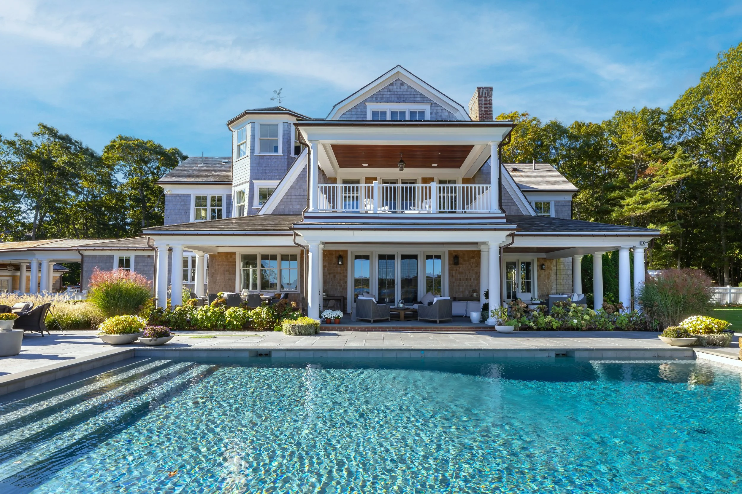 Luxury Cape Cod home with pool, covered balconies, white columns, and landscaped outdoor living areas.