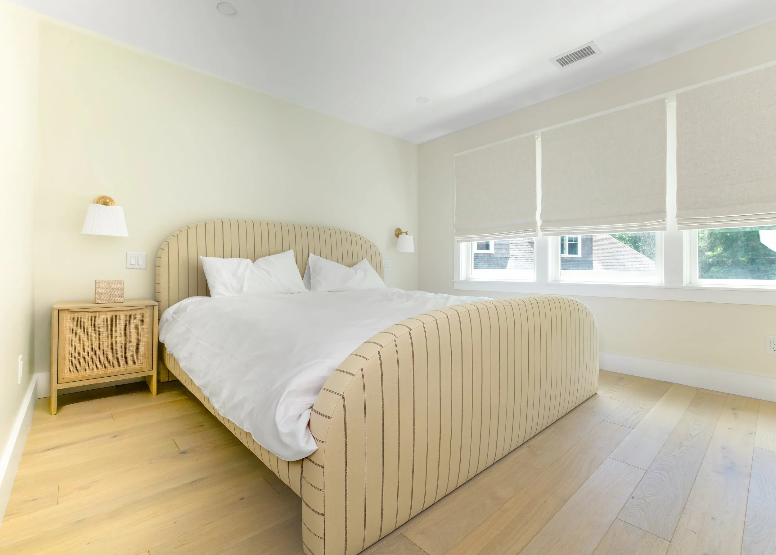 Custom home bedroom featuring light hardwood floors, upholstered furniture, large windows, soft neutral finishes, and luxury Cape Cod residential interior design.