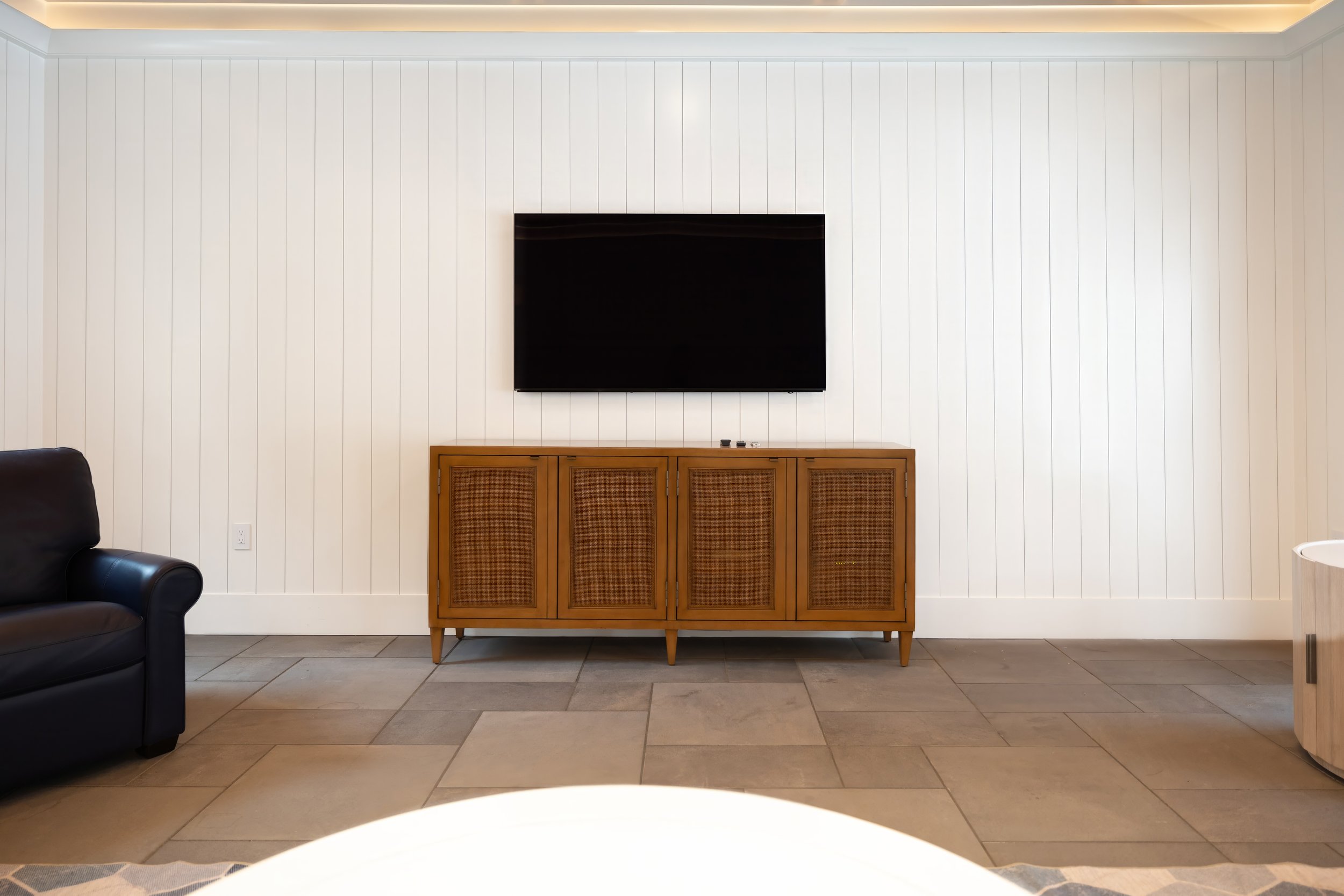 Pool house living area with white paneled walls, stone flooring, wall-mounted TV, and wood cabinet.