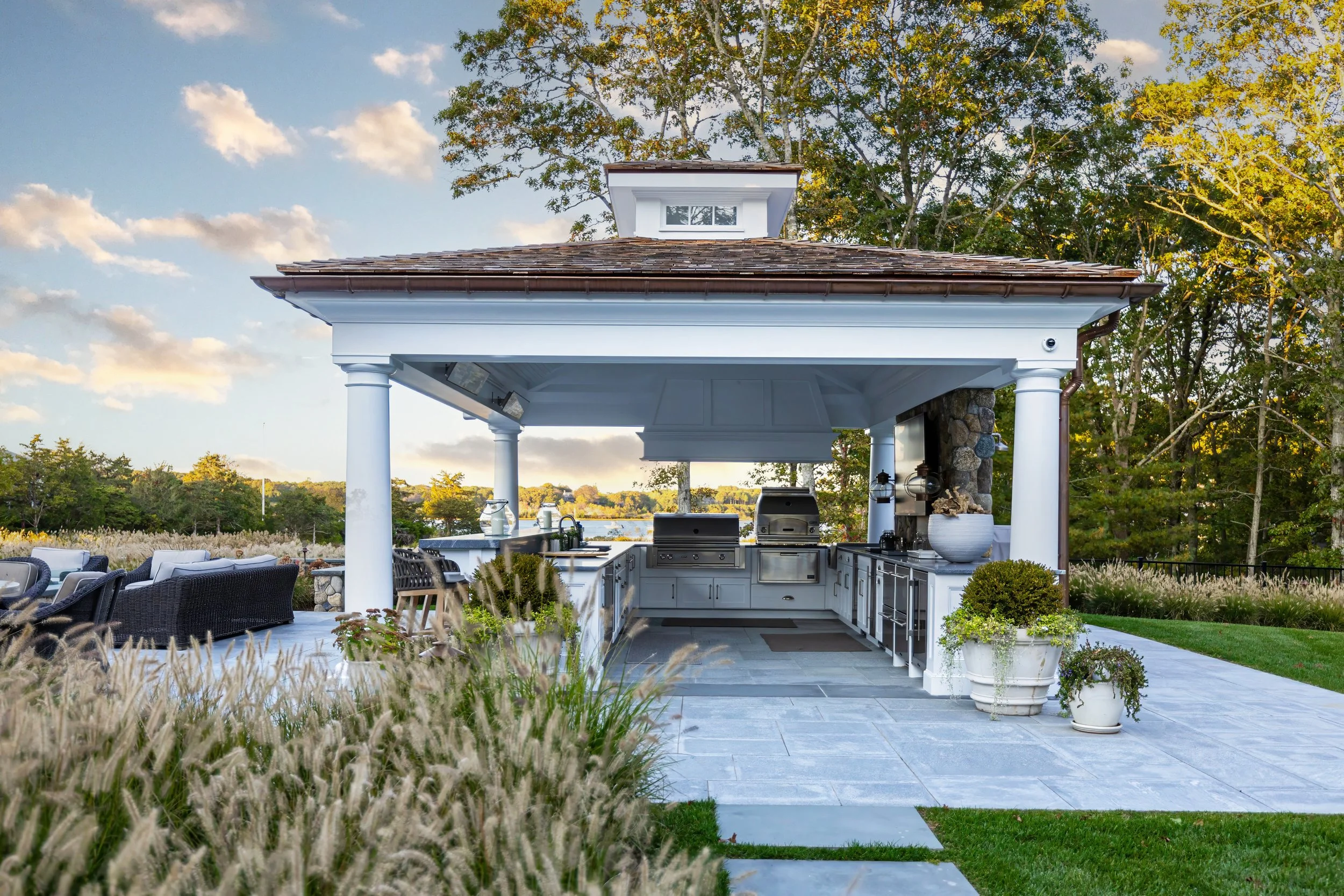 Covered outdoor kitchen with white columns, stone patio, ornamental grasses, and water views on Cape Cod.