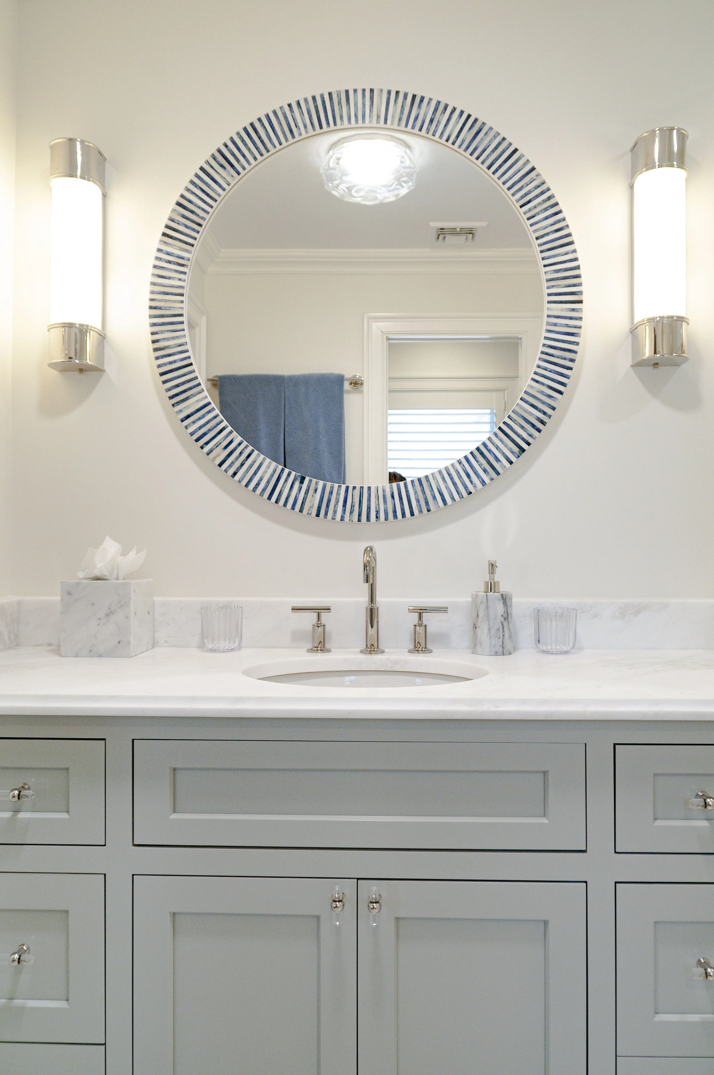 Luxury Cape Cod bathroom vanity with blue-trimmed round mirror, marble counter, polished sconces, and custom cabinetry.