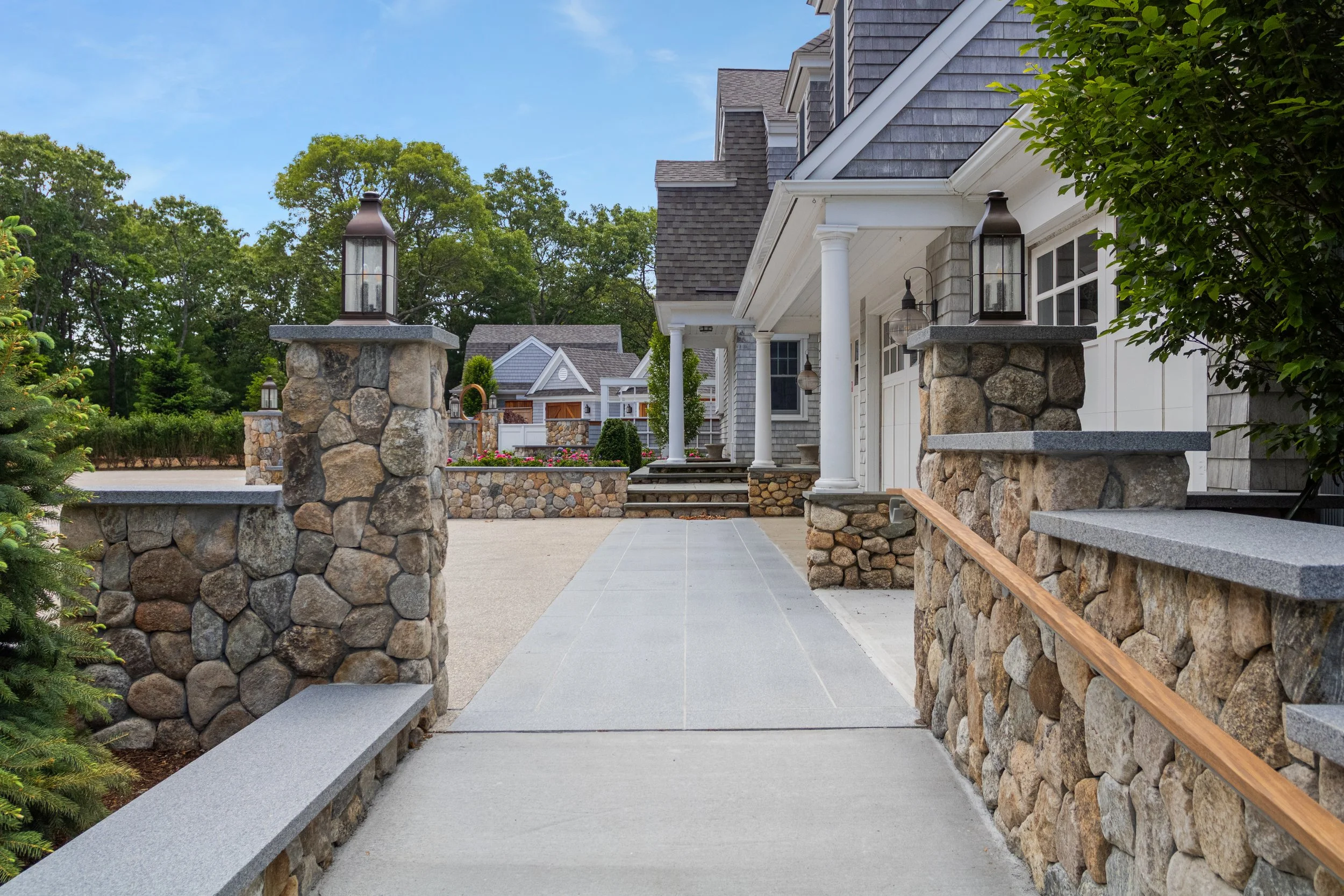 Stone-lined walkway with lantern pillars leading to a shingle-style Cape Cod home with white columns.