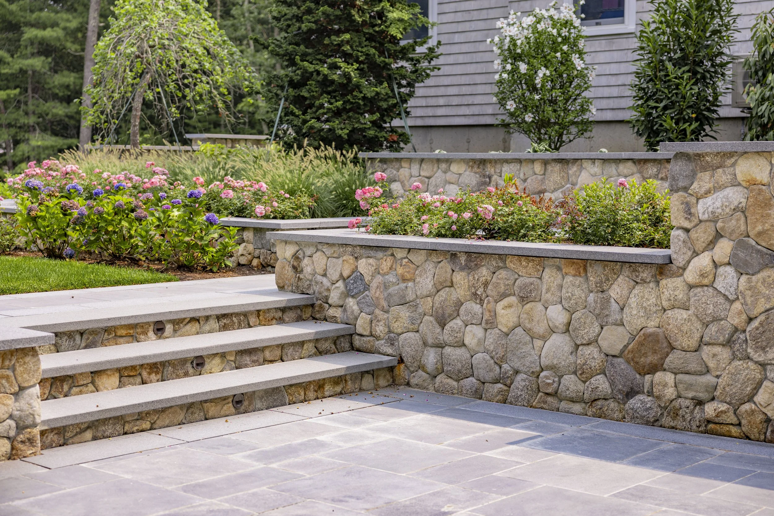 Cape Cod home garden with stone walls, slate patio, steps, and flowering landscape beds.