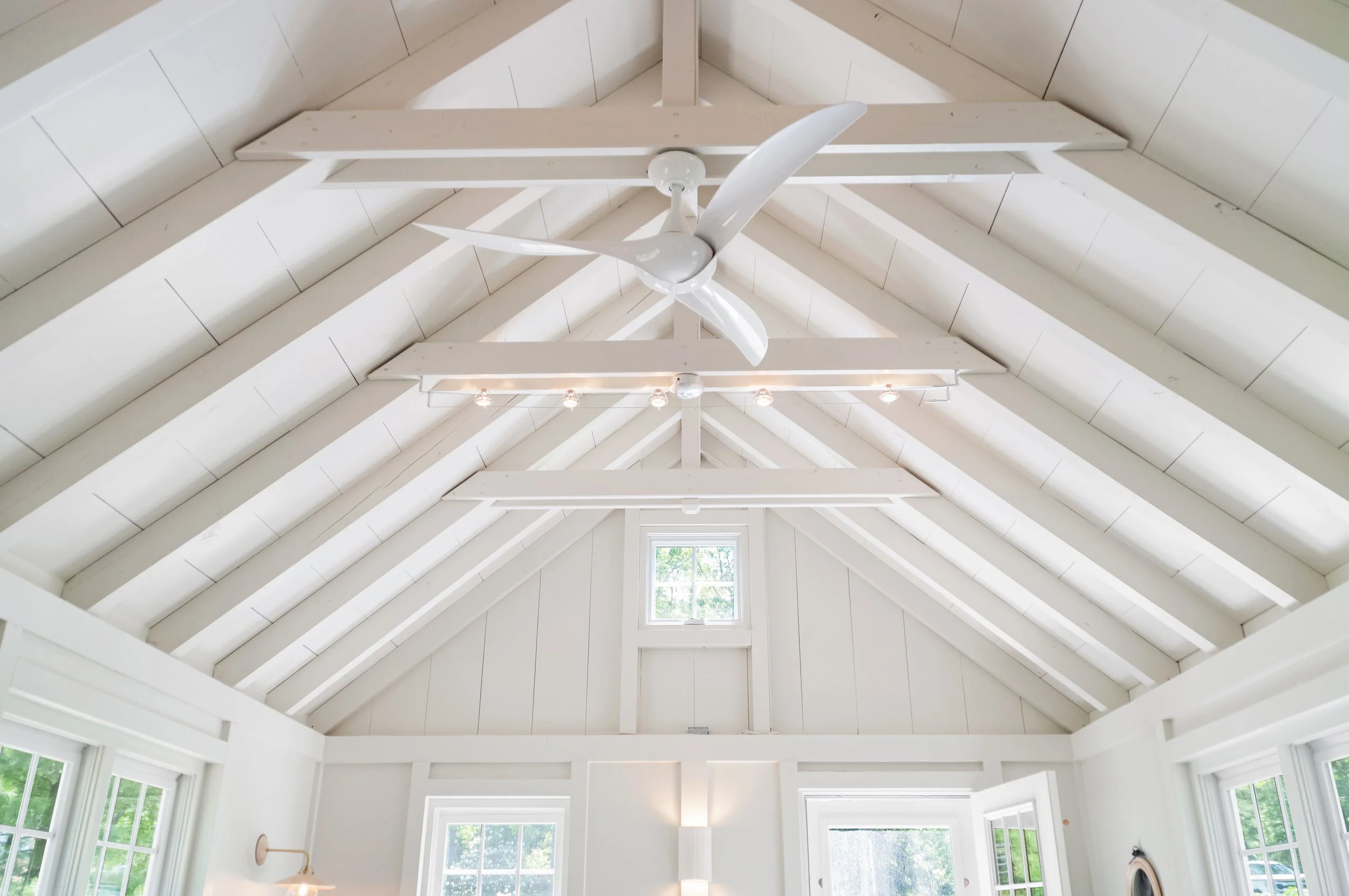White vaulted ceiling with exposed rafters, track lighting, and a ceiling fan in a coastal living space.