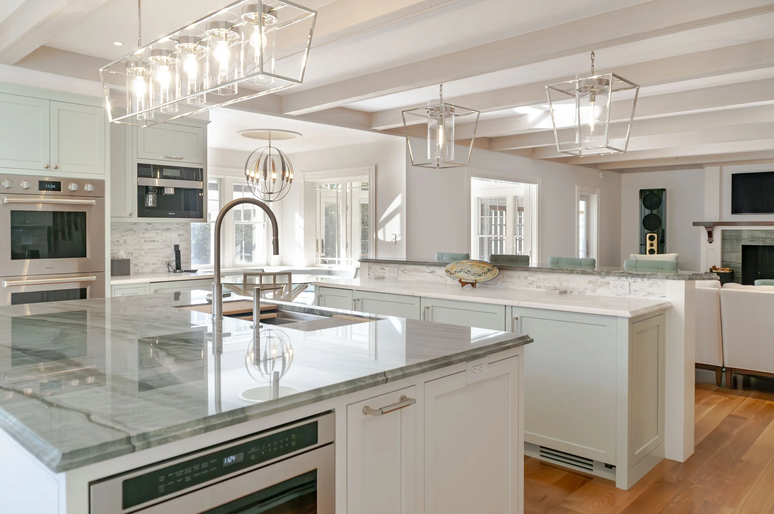 Sunlit custom coastal home kitchen with marble countertops, pale cabinetry, statement lighting, and open living area.
