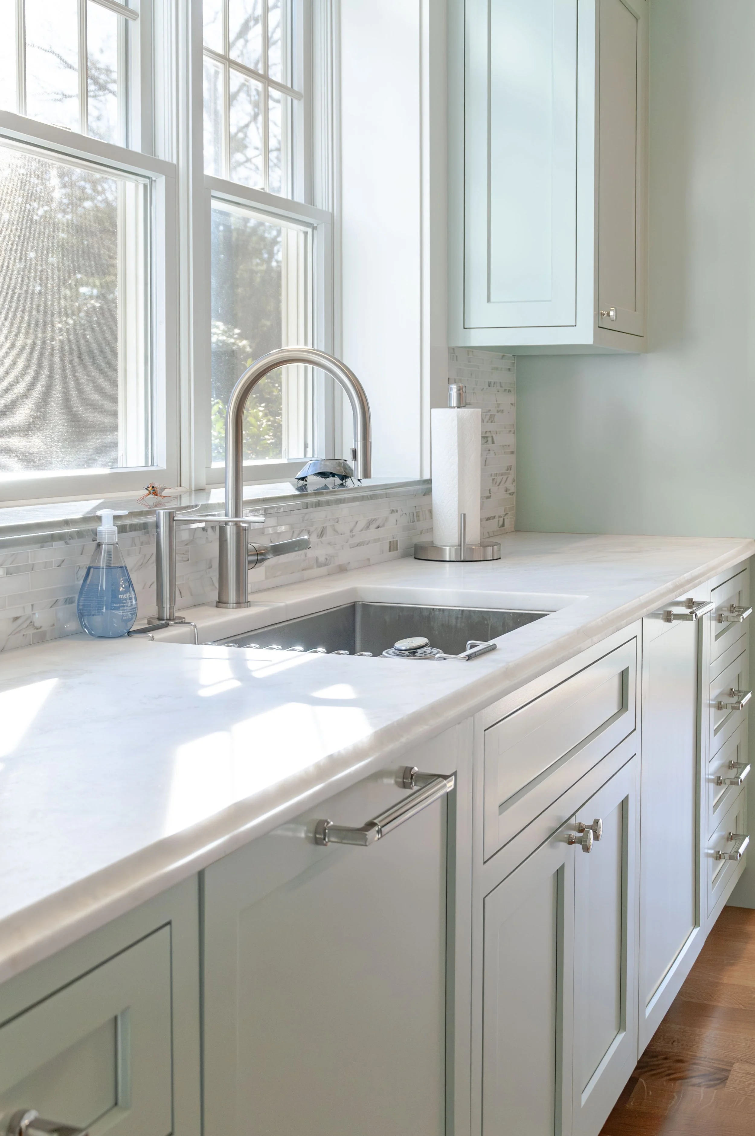 Cape Cod kitchen sink with tall windows, marble counters, pale cabinetry, and polished fixtures.